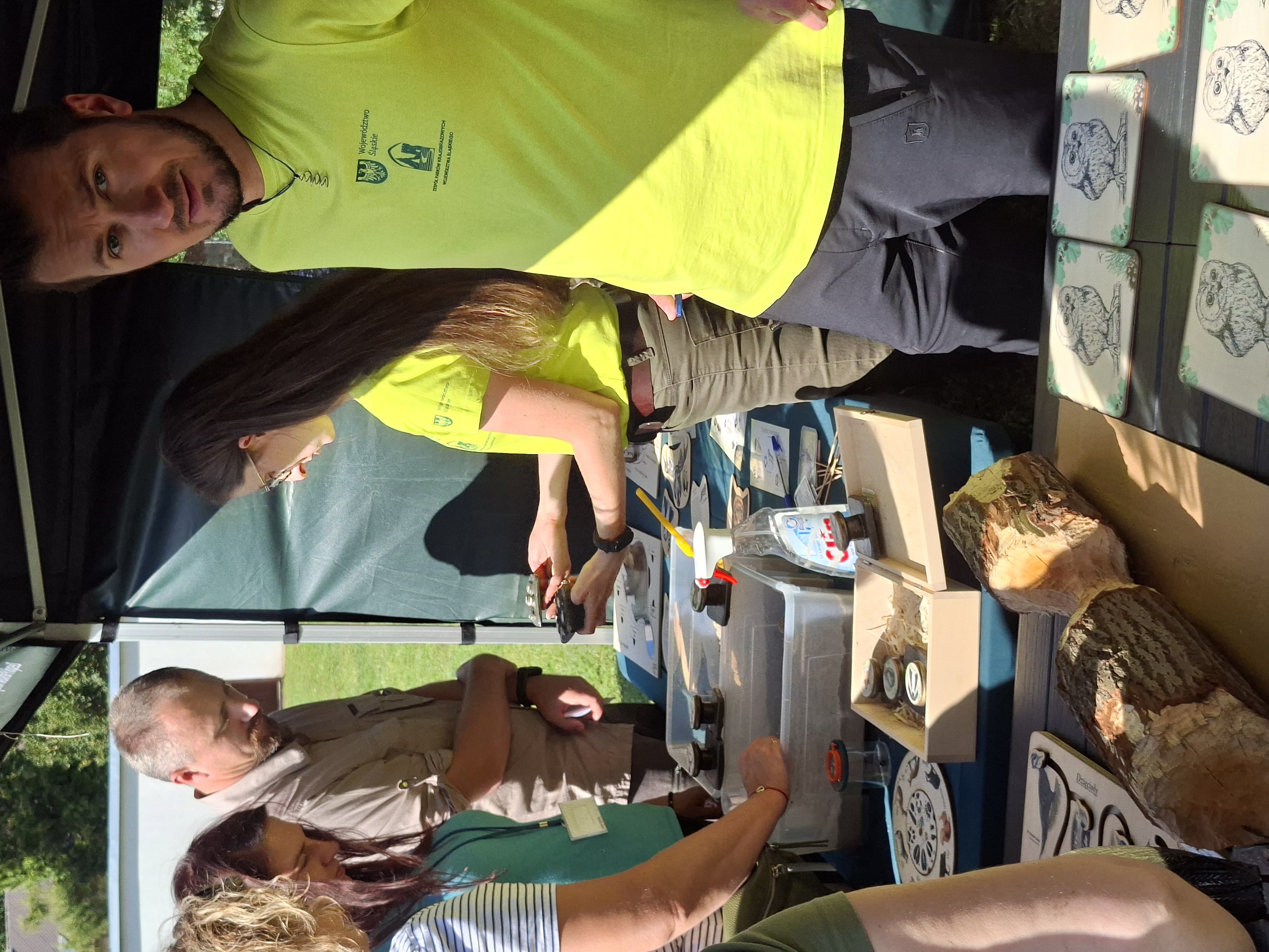 Z prawej części widoczny mężczyzna z ciemnym zarostem na twarzy, w jasnozielonej koszulce stojący przy stole, na którym znajdują się pomoce dydaktyczne - tropy zwierząt do odciskania w piasku. Za mężczyzną widoczna kobieta z długimi włosami ubrana w jasnozieloną koszulkę, która pokazuje coś osobom stojącym po drugiej stronie stołu.