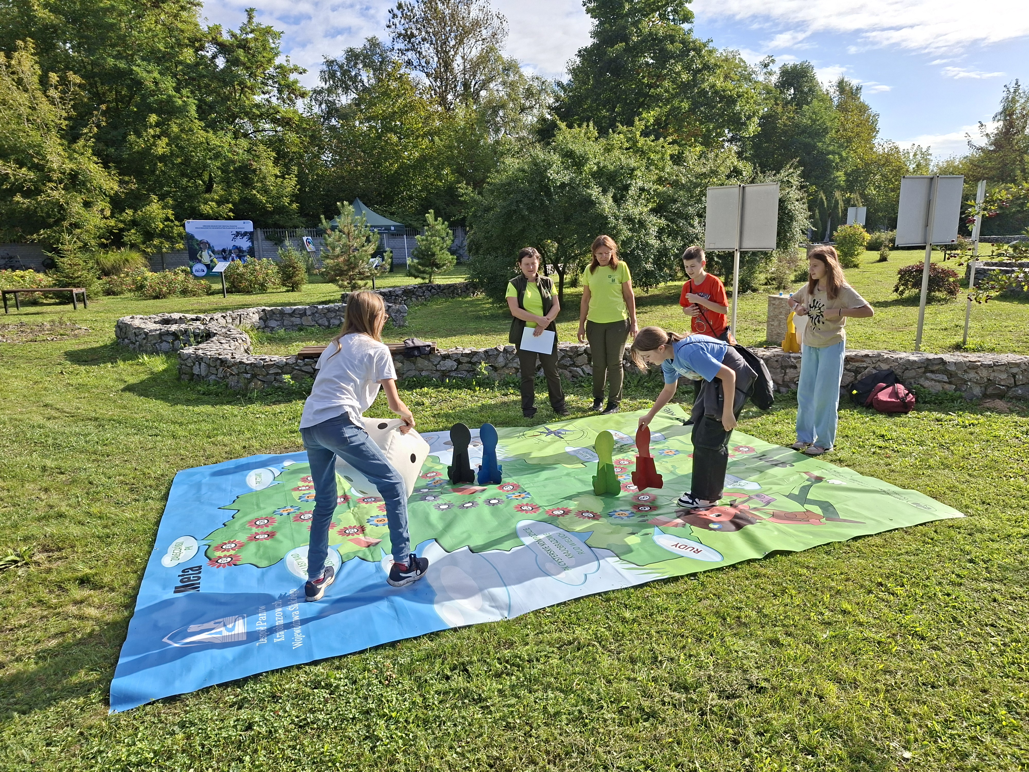 Na trawniku rozłożona jest duża, kolorowa plansza do gry, przedstawiająca kontur województwa śląskiego. Na planszy stoi dziewczyna z długimi włosami, ubrana w jasną koszulkę i niebieskie spodnie, która trzyma w dłoniach dużą, biało-czarną kostkę do gry. Po przeciwnej stronie stoi pochylona do przodu dziewczyna w niebieskiej koszulce, która ustawia duży, czerwony pionek do gry. W tle stoją cztery osoby obserwujące grę.