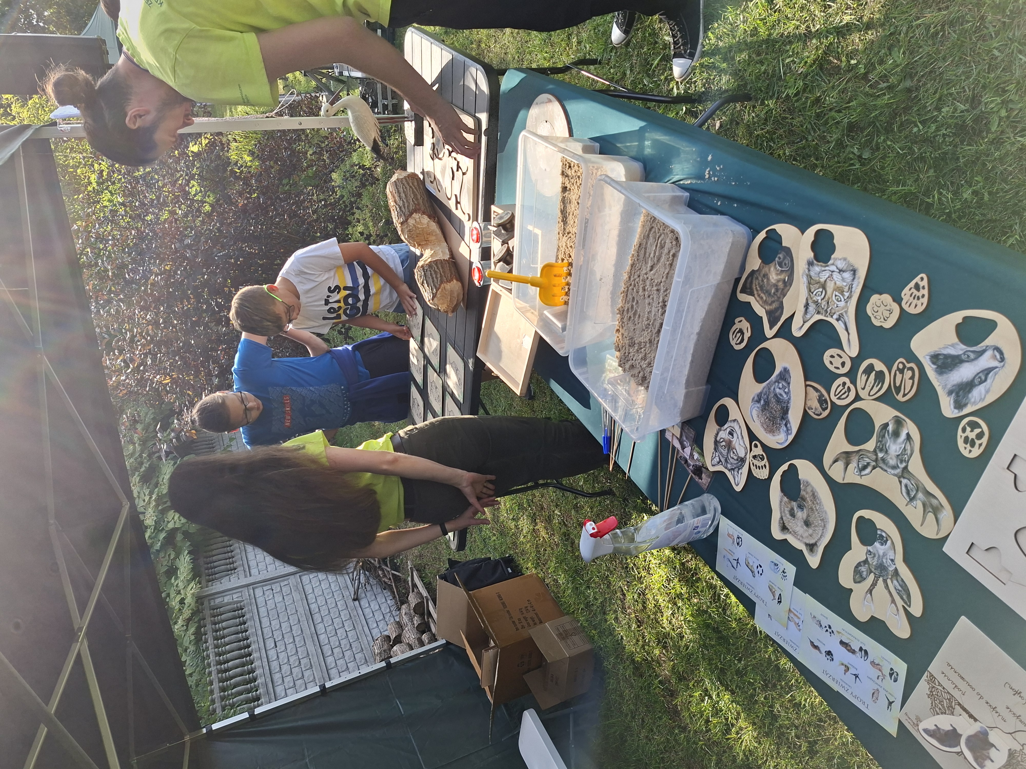 Na pierwszym planie widoczny blat stołu z zielonym obrusem, na którym znajdują się pomoce dydaktyczne - puzzle, piasek i tropy zwierząt do odciskania w piasku. W tle kobieta z długimi włosami i jasnozielonej koszulce obserwuje dwóch chłopców, którzy układają karty memory. Całe stoisko znajduje się w zielonym namiocie.