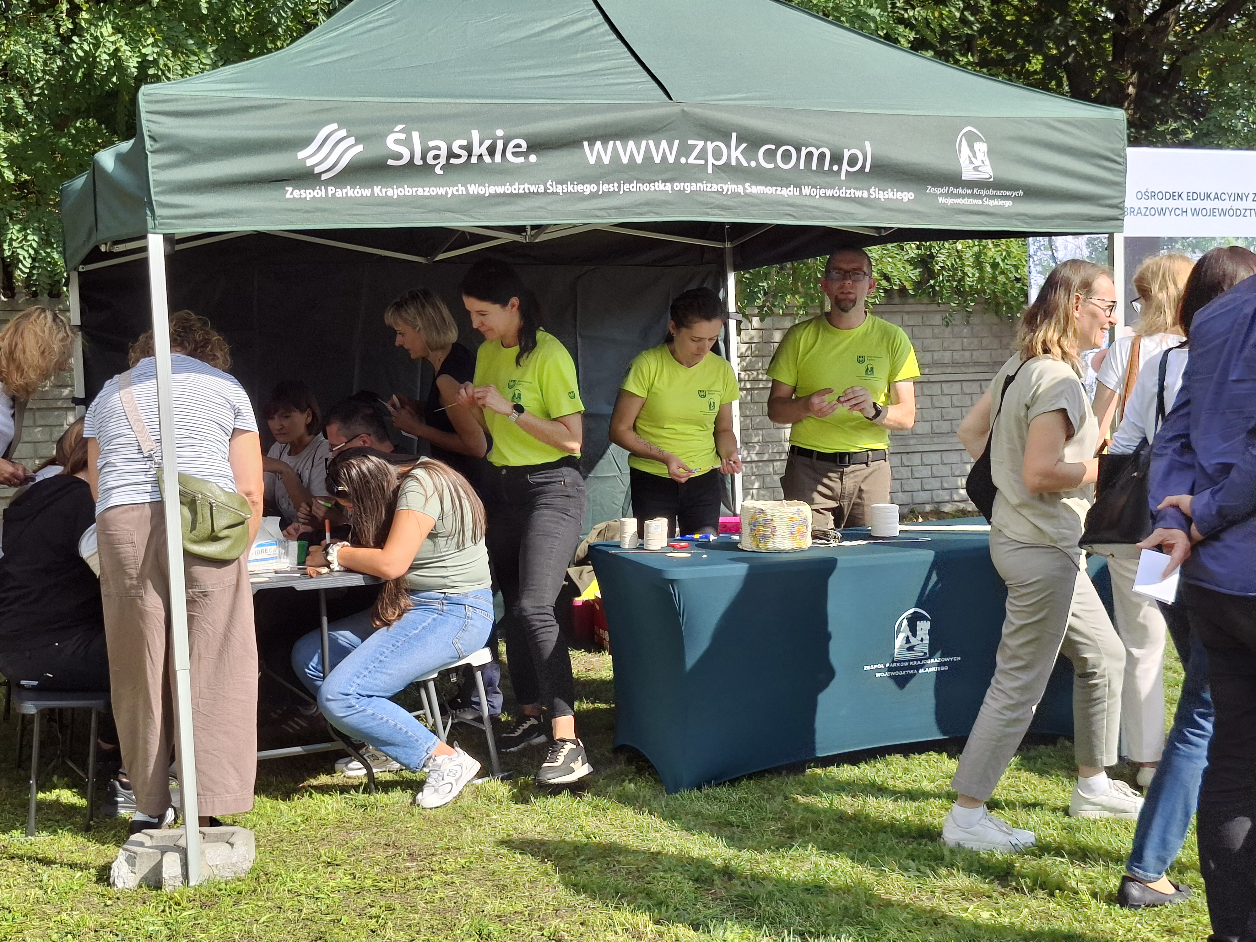 Na zdjęciu widoczny zielony namiot, pod którym ustawione są dwa stoły. Na stole nakrytym zielonym obrusem znajdują się materiały plastyczne. Przy stole stoją dwie osoby ubrane w jasnozielone koszulki - mężczyzna w okularach i kobieta z ciemnymi włosami. Z lewej strony widoczny stół, przy którym siedzi grupa kobiet, wykonujących prace plastyczne.