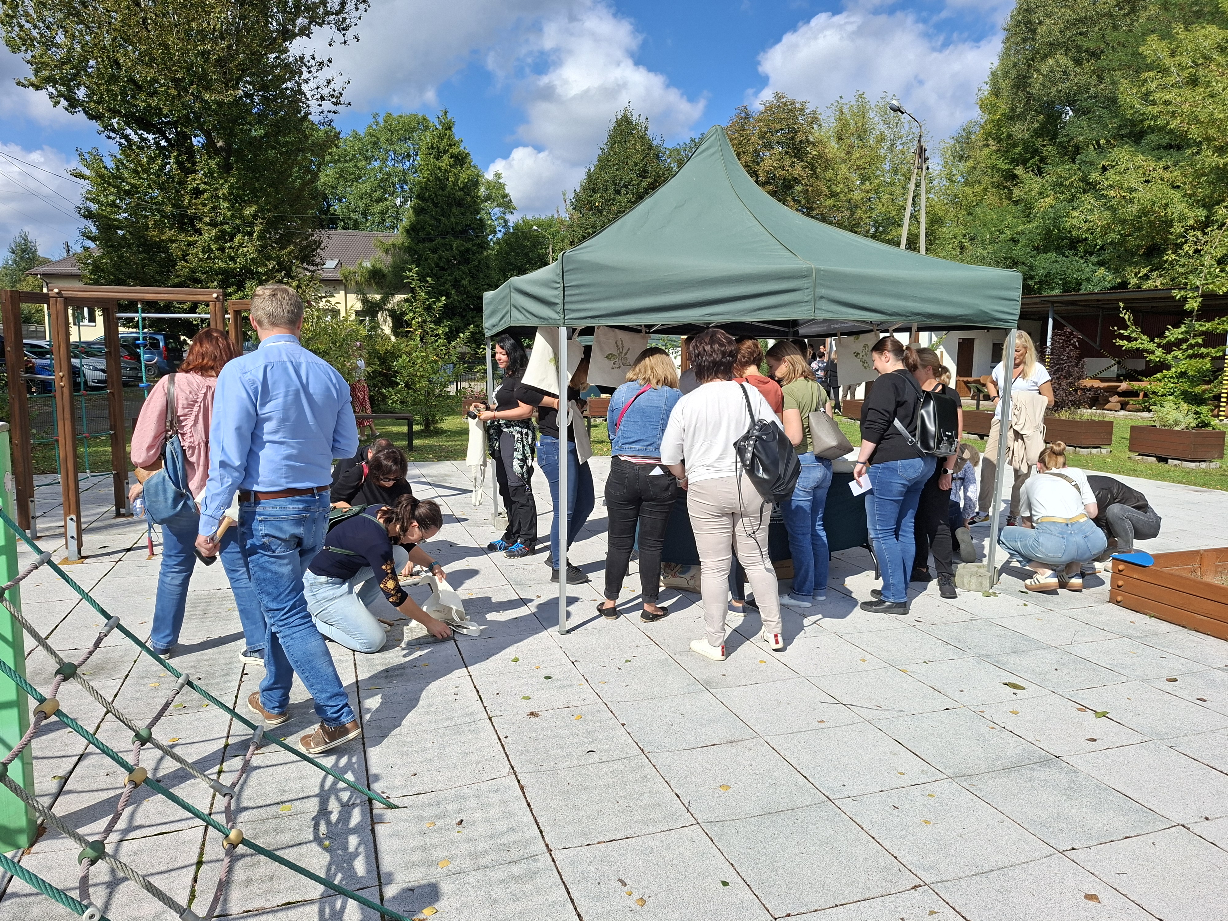 Na zdjęciu widoczny fragment placu zabaw z miękkim podłożem. W lewej części zdjęcia widoczny mężczyzna ubrany w niebieskie ubranie, stojący tyłem do fotografa. Mężczyzna obserwuje osoby, które w pozycji klęczącej wykonują prace rękodzielnicze na podłożu. Z lewej strony widoczny zielony namiot, pod którym gromadzi się grupa osób zainteresowanych wykonywaniem prac rękodzielniczych.