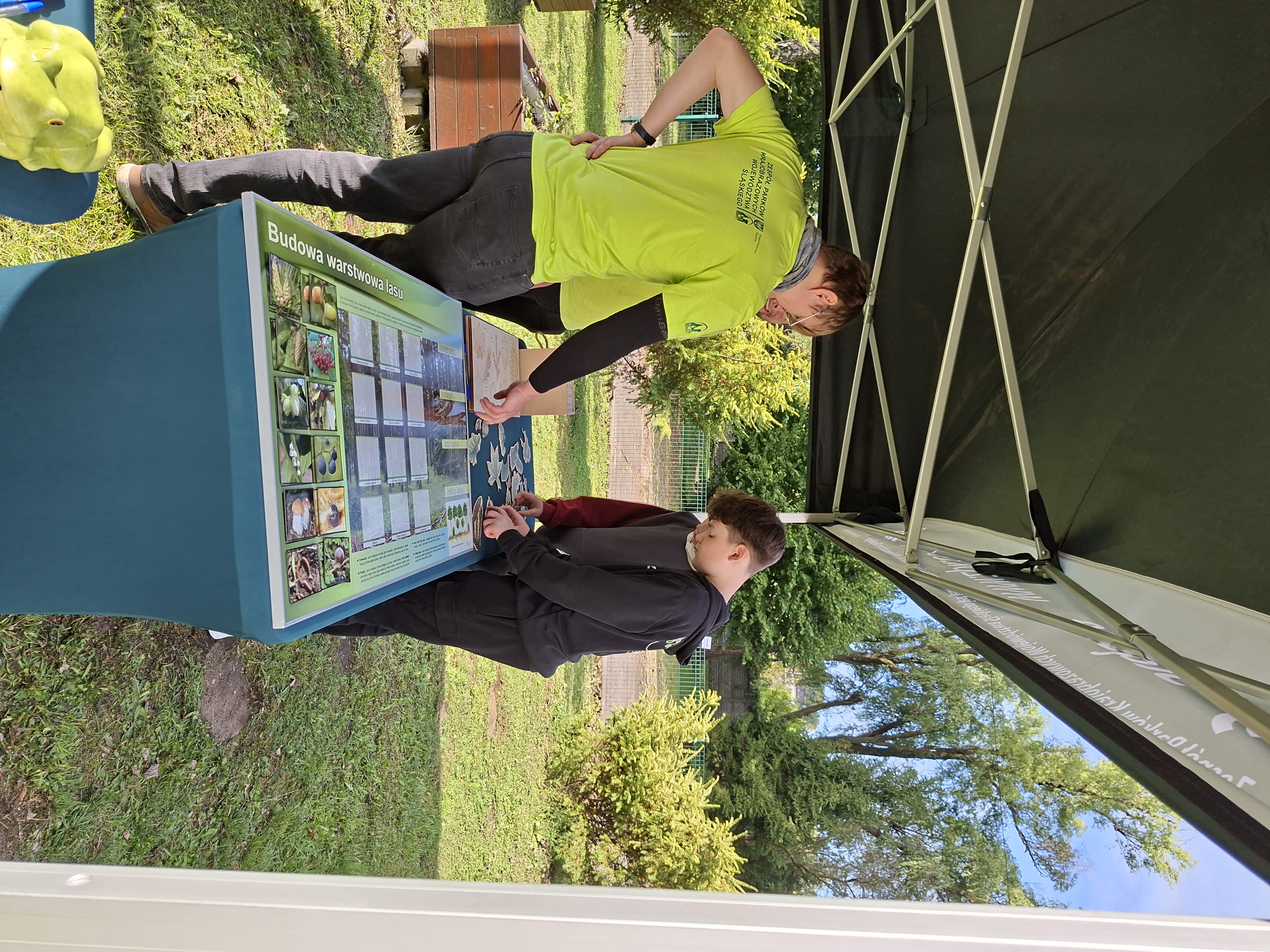 Na zdjęciu mężczyzna w okularach, ubrany w jasnozieloną koszulkę stojący przy stole z pomocami dydaktycznymi. Mężczyzna wskazuje prawą ręką na jeden z elementów rozłożonej na stole układanki. Po przeciwnej stronie stołu stoi chłopiec w czarnej bluzie z kapturem. Chłopiec zajęty jest układaniem elementów układanki. Stół ustawiony jest pod zielonym namiotem.