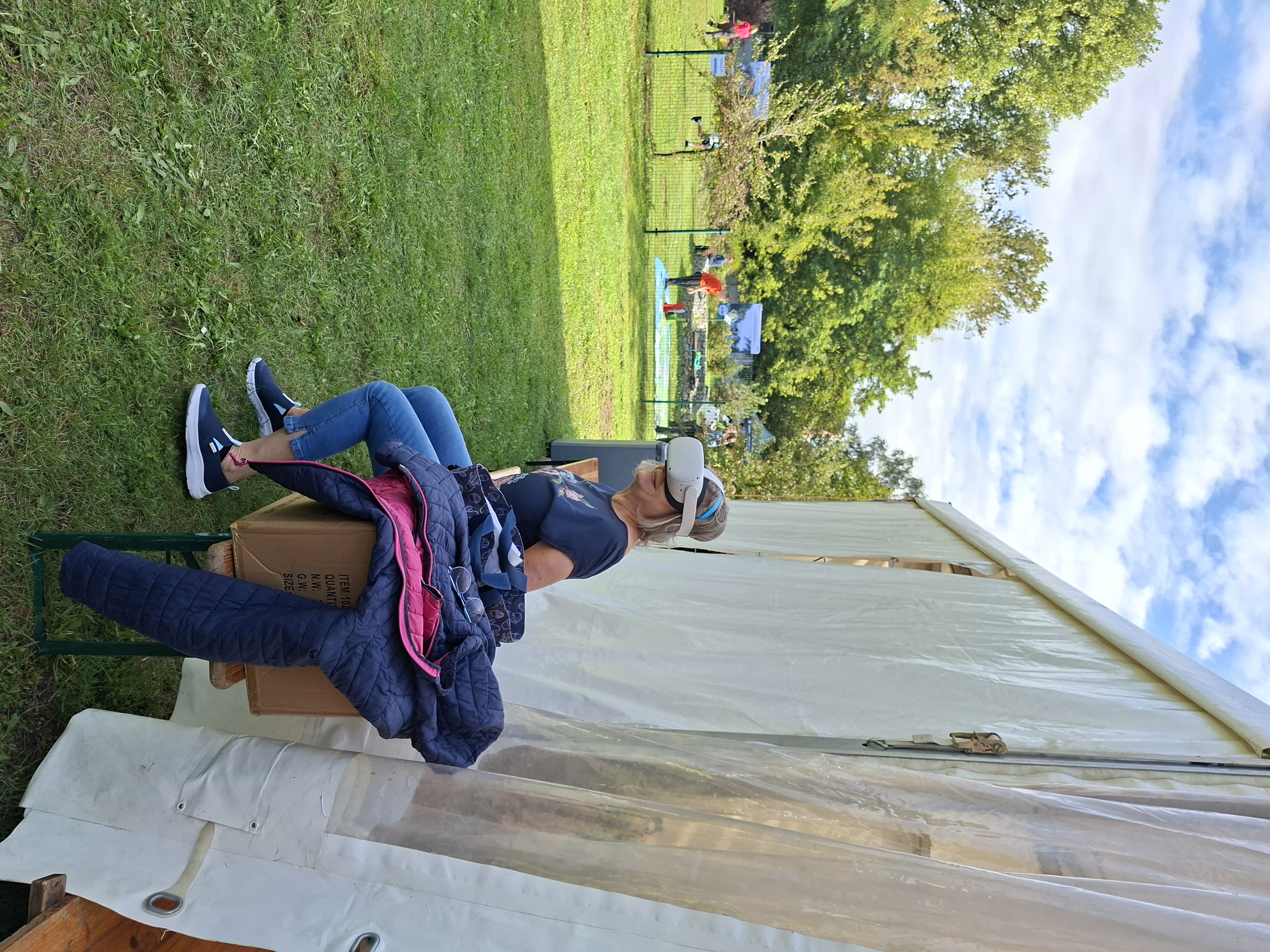 Na zdjęciu widoczna kobieta z blond włosami, ubrana w granatową bluzkę i dżinsy. Na głowę założyła okulary VR, w których odtwarzany jest obraz. W tle widoczna grupa dzieci grająca w grę planszową rozłożoną na trawniku.