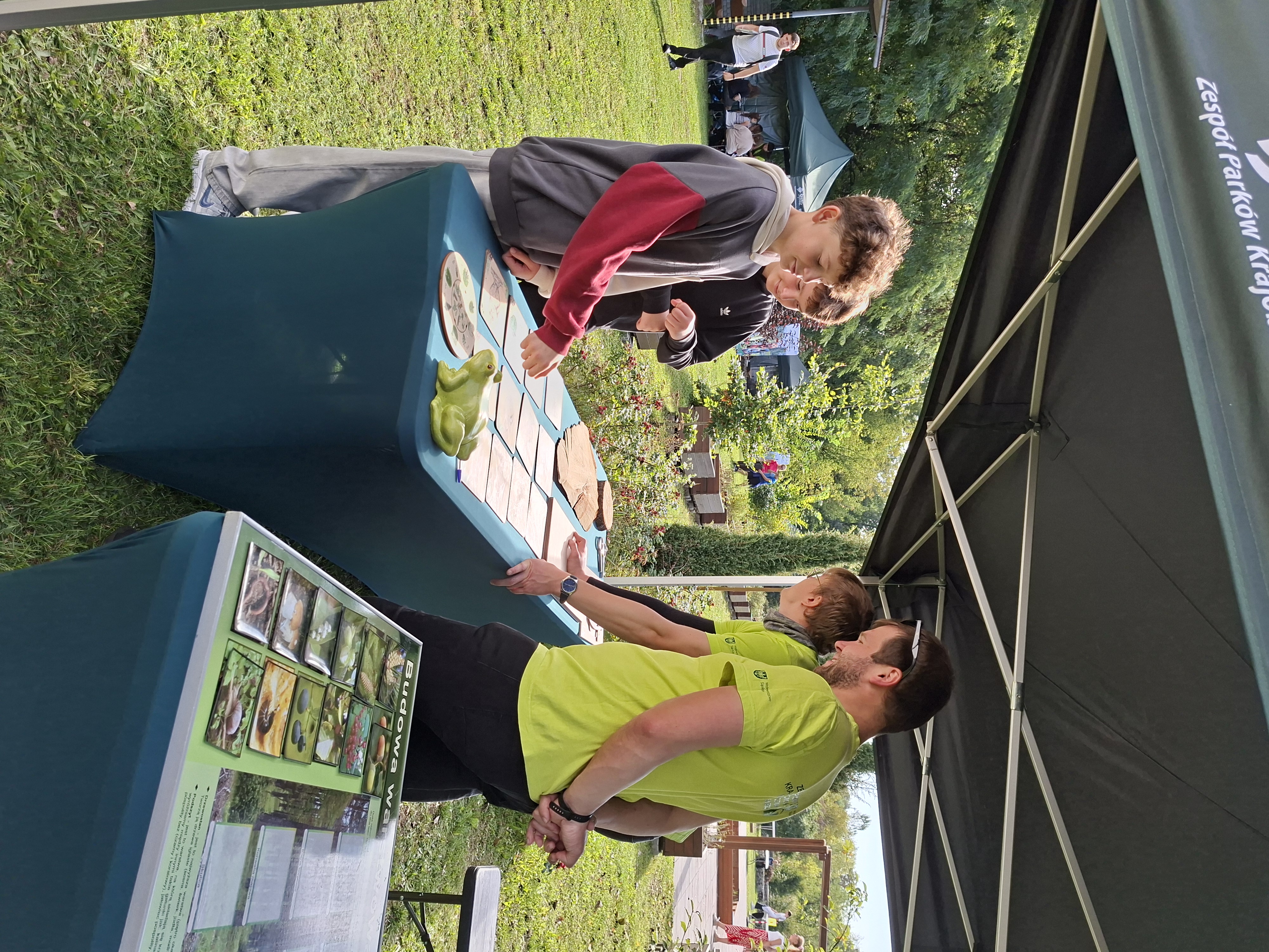 Na zdjęciu widoczni mężczyźni w jasnozielonych koszulkach, stojący bokiem do fotografującego. Przed mężczyznami stoi zielony stół, na którym leżą karty do gry w memory z obrazkami zwierząt, wykonane ze sklejki drewnianej. Po przeciwnej stronie stołu stoją ubrani w sportowe bluzy chłopcy, którzy grają w karty memory.