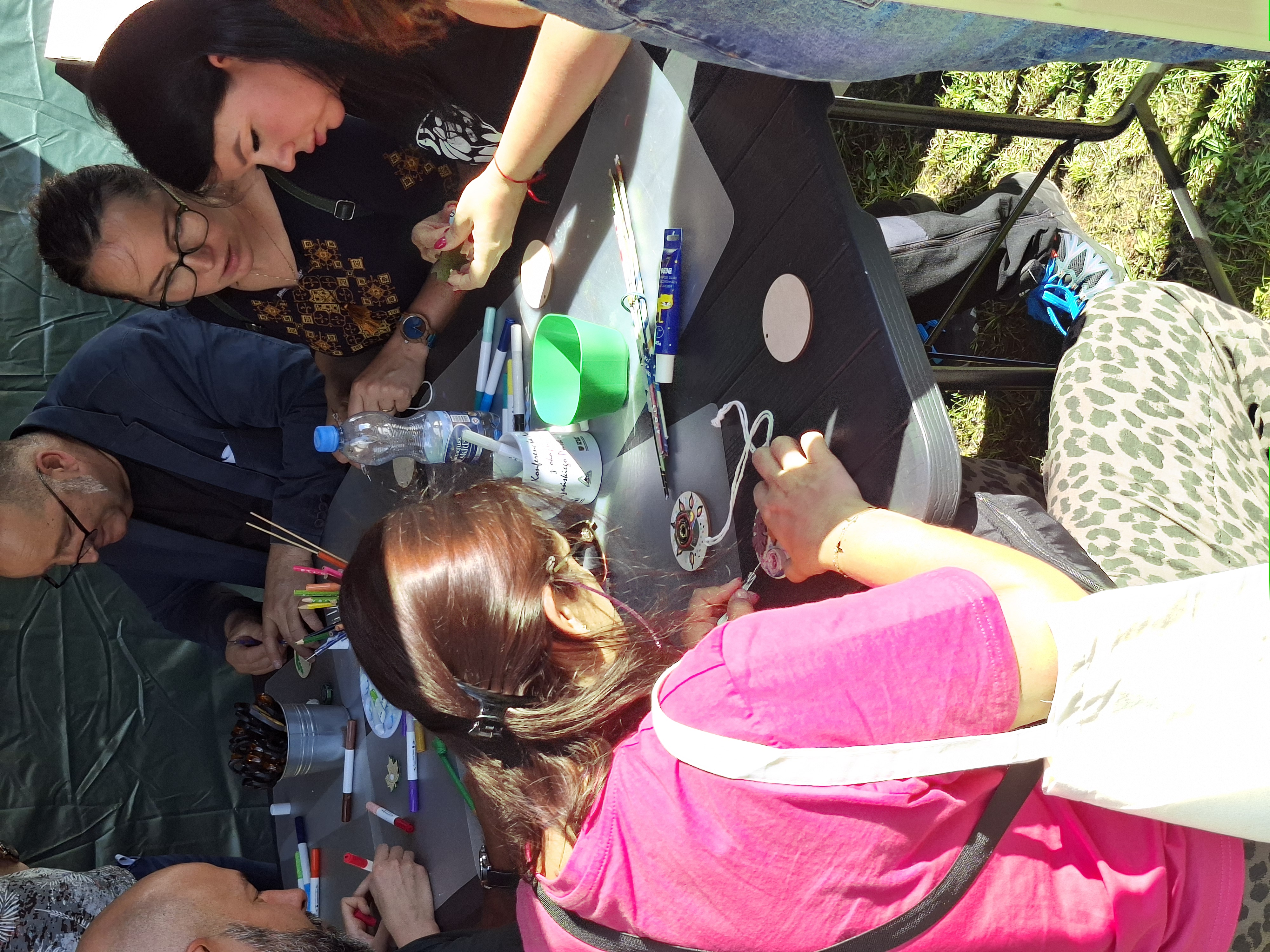 Na pierwszym planie ciemnowłosa kobieta w różowej bluzce, siedząca przy stole, wykonuje prace rękodzielnicze. Po przeciwnej stronie stoły mężczyzna w okularach i granatowej marynarce oraz dwie ciemnowłose kobiety w ciemnych bluzkach, wykonują prace rękodzielnicze. Na stole znajdują się różne materiały: flamastry, pędzelki, kubeczki, klej, drewniane krążki, sznurek.