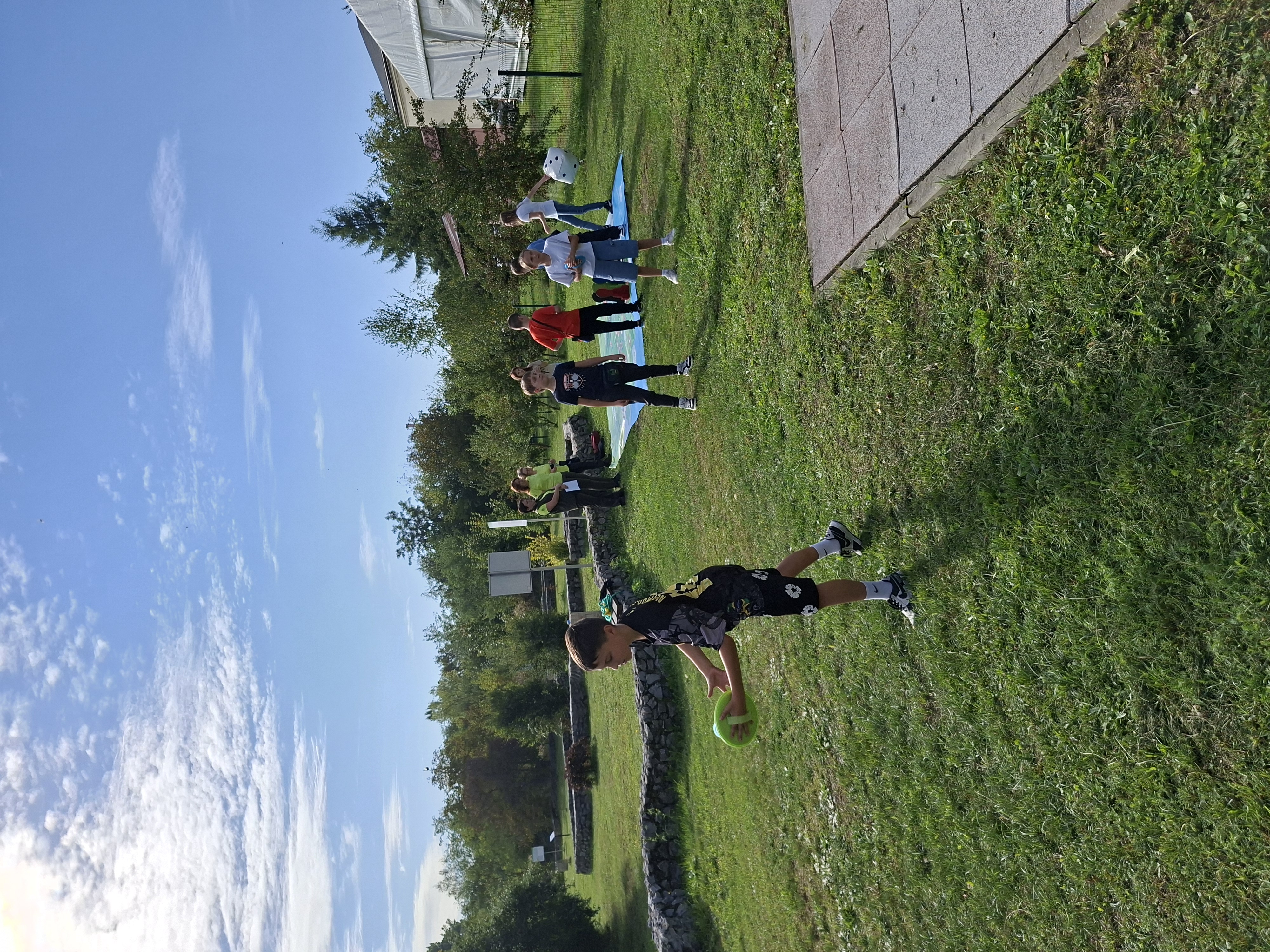 Na pierwszym planie chłopiec w ciemnym ubraniu, trzymający w lewej dłoni piłeczkę tenisową. W tle grupa dzieci grająca w dużą grę plenerową (plansza rozłożona na trawniku). Dziewczynka w jasnej koszulce, trzymająca w prawej dłoni dużą kostkę do gry, przygotowuje się do rzutu. W tle zielone drzewa i krzewy oraz błękitne niebo.