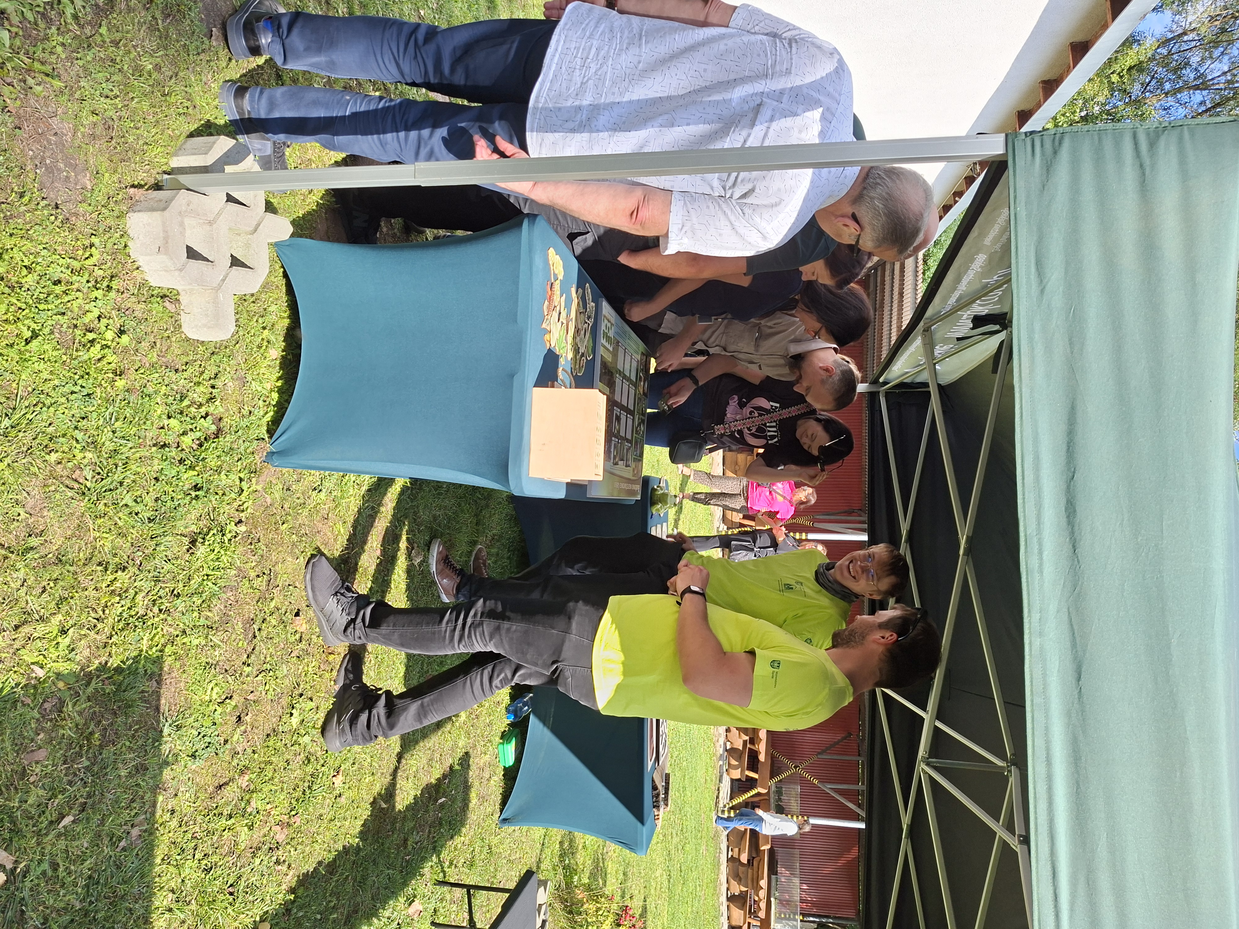 Na pierwszym planie (z lewej strony) stoi szpakowaty mężczyzna w jasnej koszuli. Na zdjęciu widoczny namiot, pod którym ustawiony jest zielony stół, na którym znajdują się pomoce dydaktyczne i tablice edukacyjne. Z prawej strony stołu stoją mężczyźni w jasnozielonych koszulkach. Po przeciwnej stronie stołu stoi grupa ok. sześciu osób (kobiet i mężczyzn), które rozwiązują zadania na tablicy edukacyjnej.