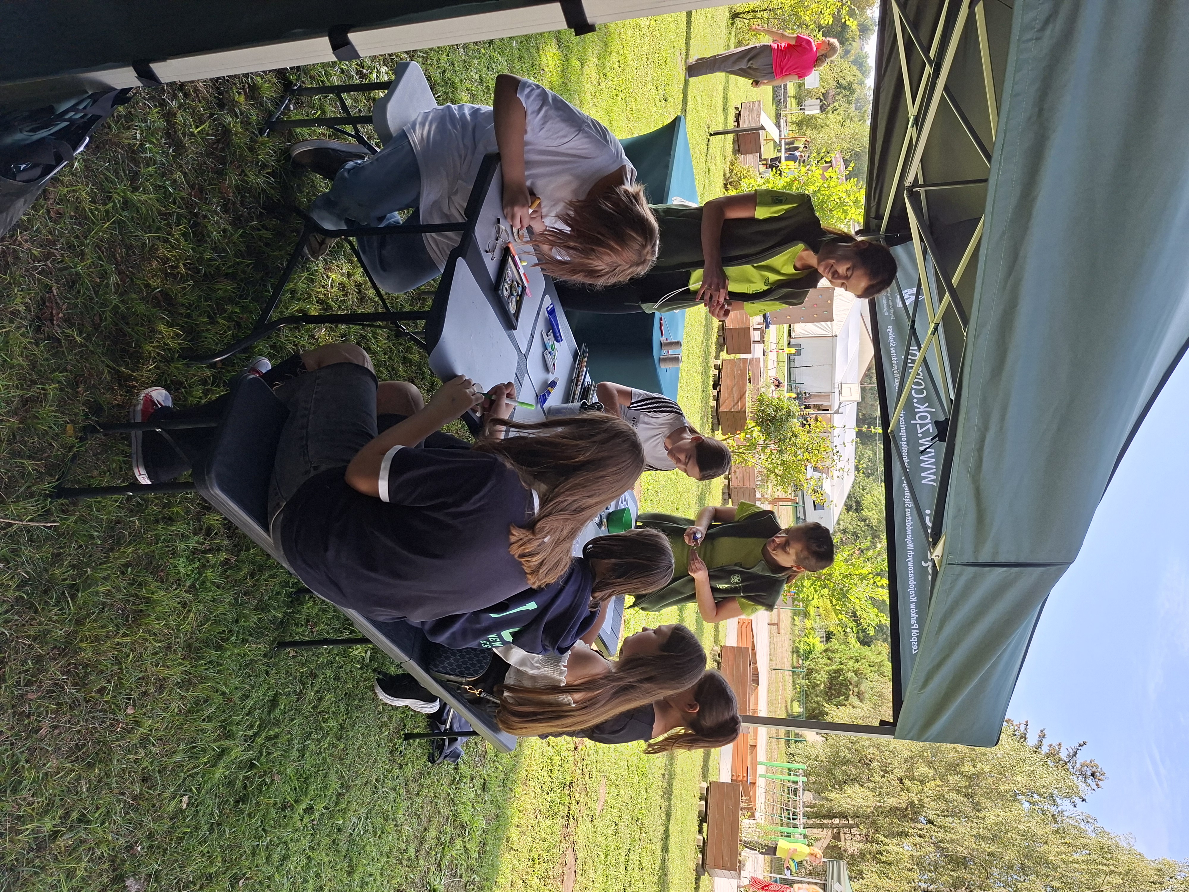 Na zdjęciu widoczny zielony namiot, pod którym stoi czarny stół. Przy stole siedzi 6 dziewcząt, które wykonują prace plastyczne. Obok stołu stoją dwie uśmiechnięte kobiety w jasnozielonych koszulkach i zielonych kamizelkach. Kobiety podają siedzącym dziewczętom materiały plastyczne.