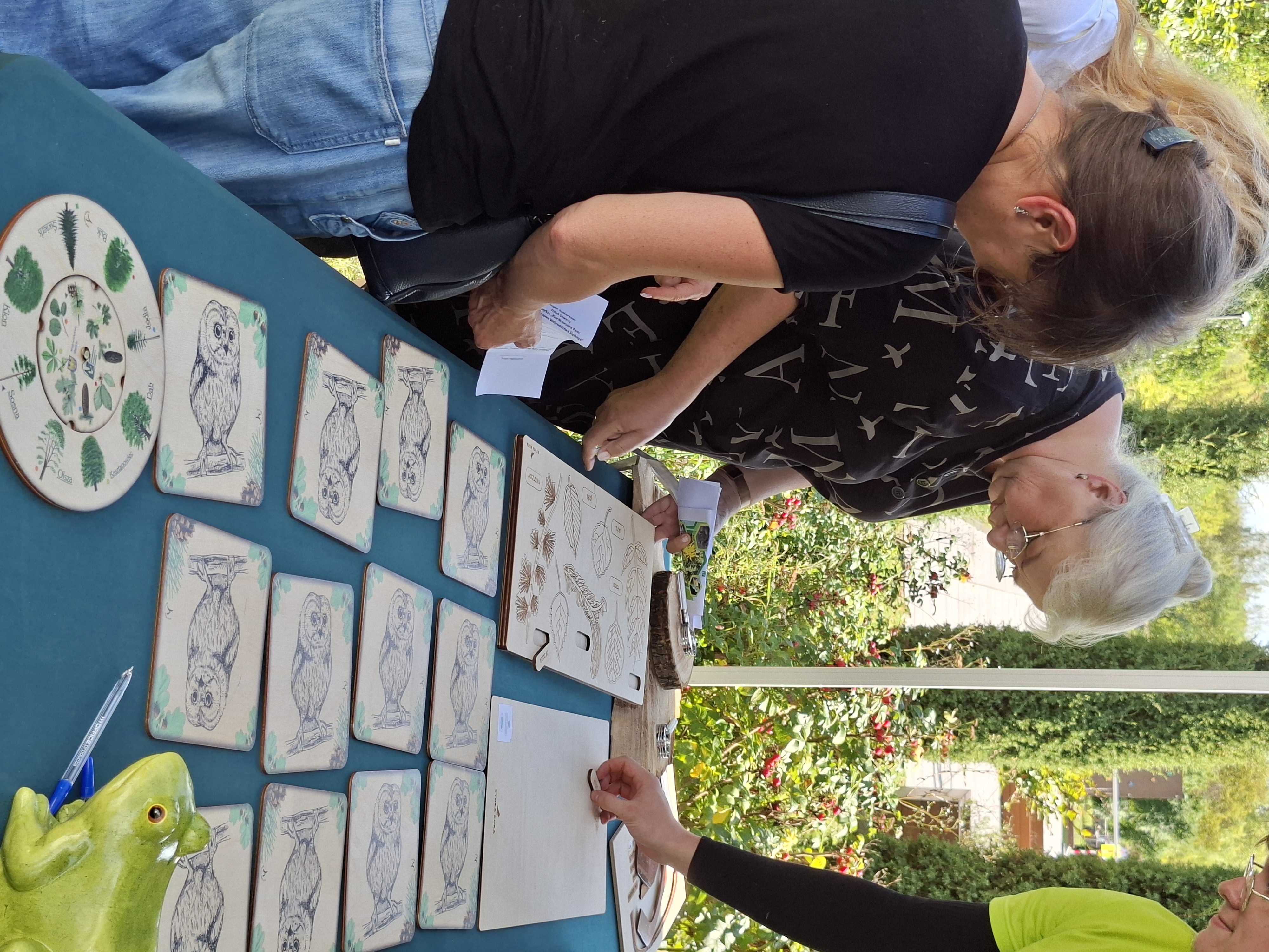 Na zdjęciu widoczne trzy kobiety w średnim wieku. Stoją przy stole na którym rozłożone są ekologiczne pomoce dydaktyczne: drewniane karty do gry memor i układanka edukacyjna polegająca na dopasowaniu liści drzew do owoców danego gatunku. Kobiety układają poszczególne elementy układanki.