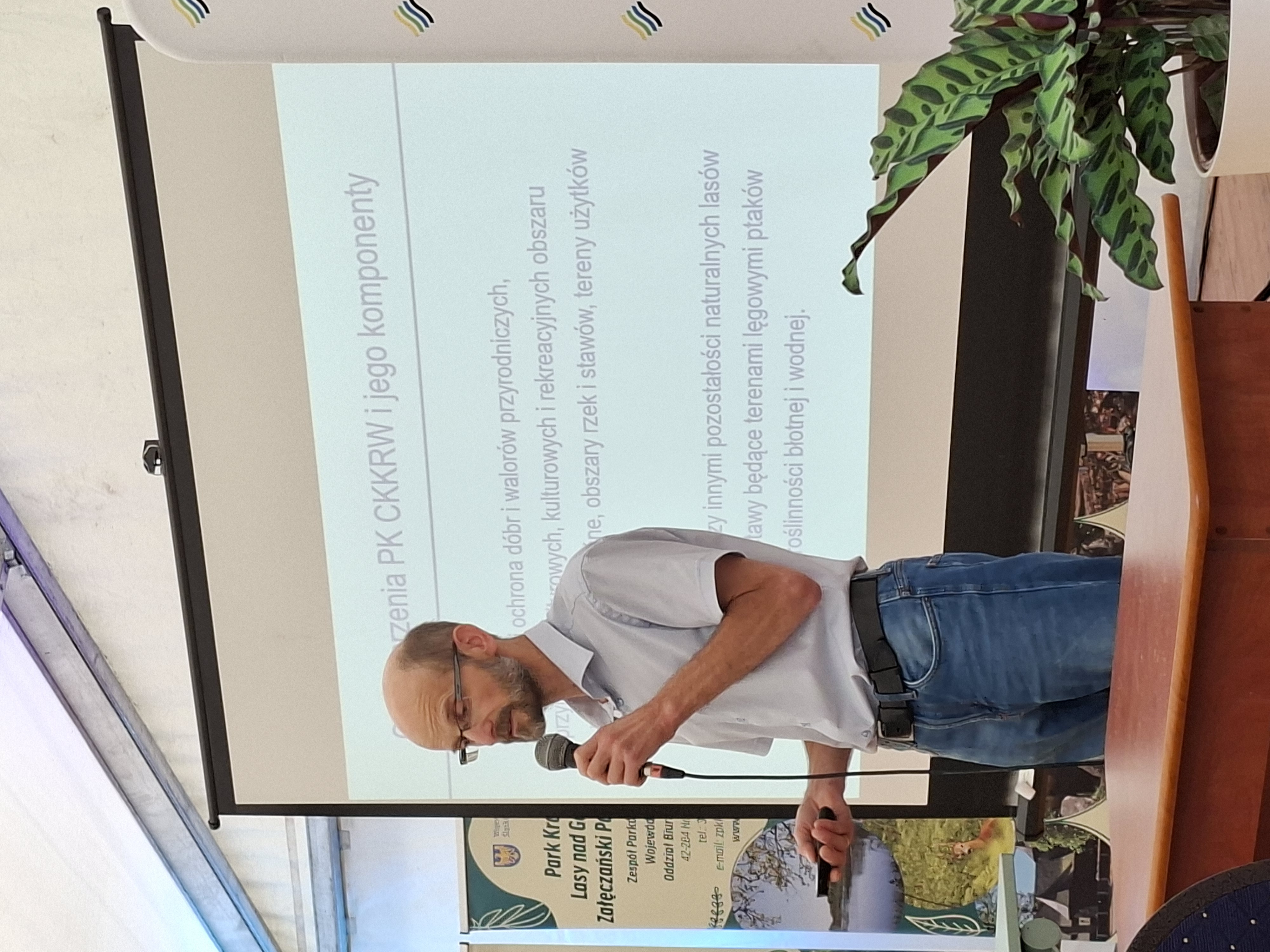 Mężczyzna w okularach i białej koszuli, stoi z mikrofonem w ręku. W tle za mężczyzną widać ekran z wyświetloną prezentacją multimedialną.