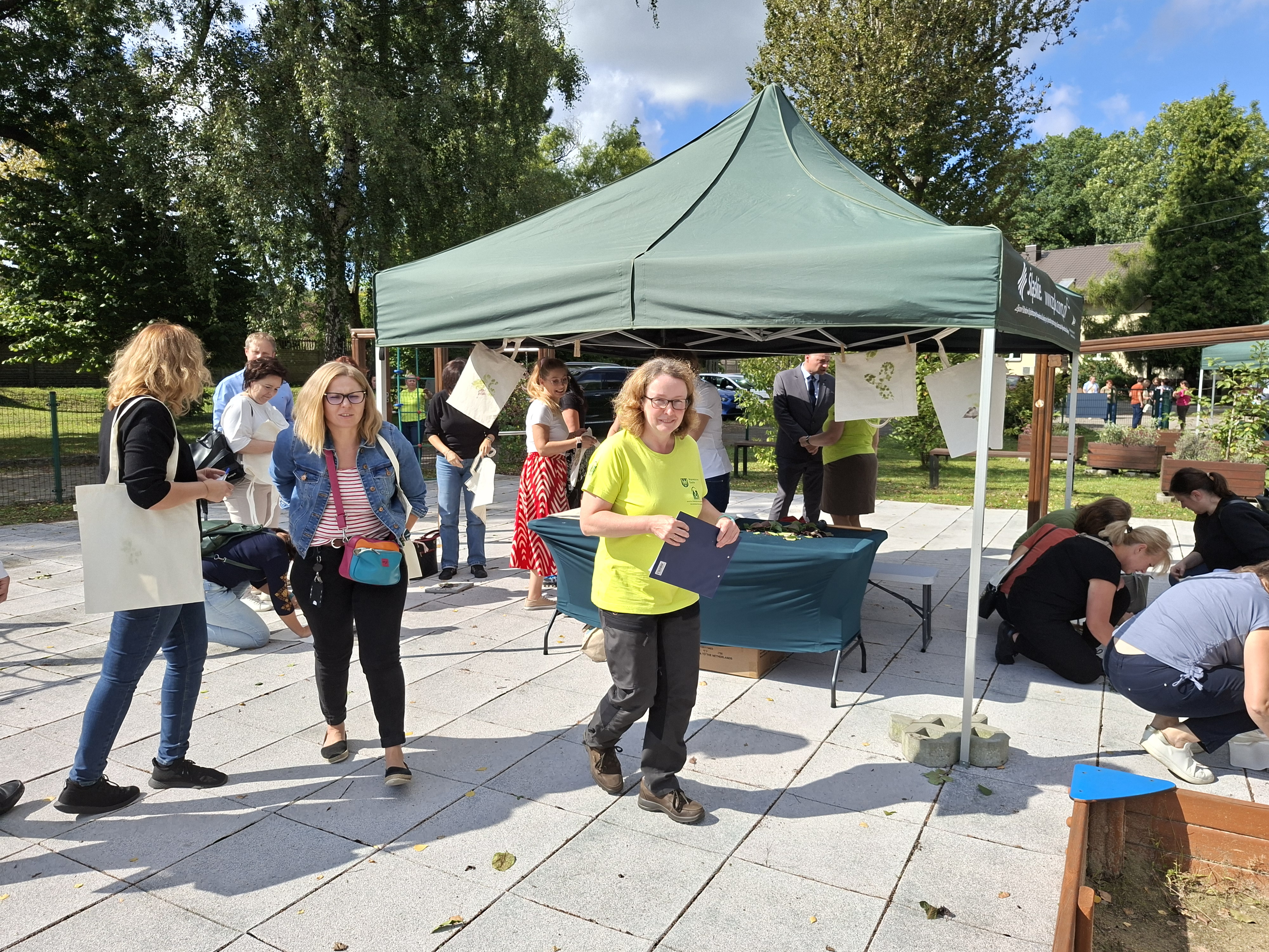 Na zdjęciu w centralnym miejscu widoczny namiot ze stoiskiem edukacyjnym. Wokół namiotu, w rozproszeniu, widoczne osoby w różnym wieku. Z lewej stronie kobiety stojące kobiety. Na pierwszym planie uśmiechnięta kobieta w jasnozielonej koszulce. Z prawej stronie grupa osób siedzi na ziemi i wykonuje prace rękodzielnicze.
