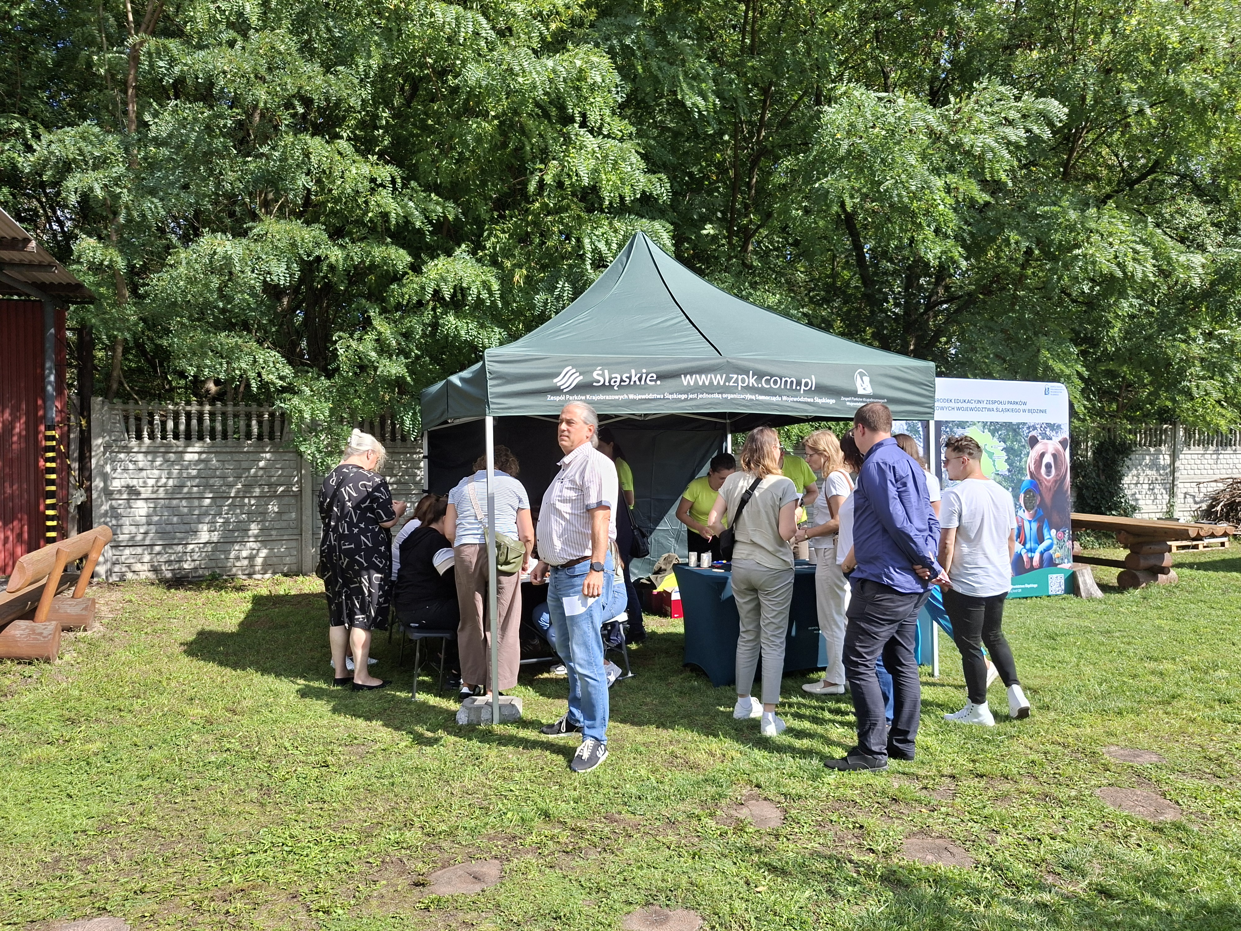 Na zdjęciu widoczny zielony namiot ze stoiskiem edukacyjnym. Przy namiocie stoi grupa osób w różnym wieku, która rozwiązuje zadania edukacyjne rozłożone na stole pod namiotem. Namiot ustawiony w plenerze. W tle zielone drzewa liściaste. Z prawej strony namiotu widoczna kolorowa ścianka do robienia zdjęć, z wyciętymi otworami na twarze.