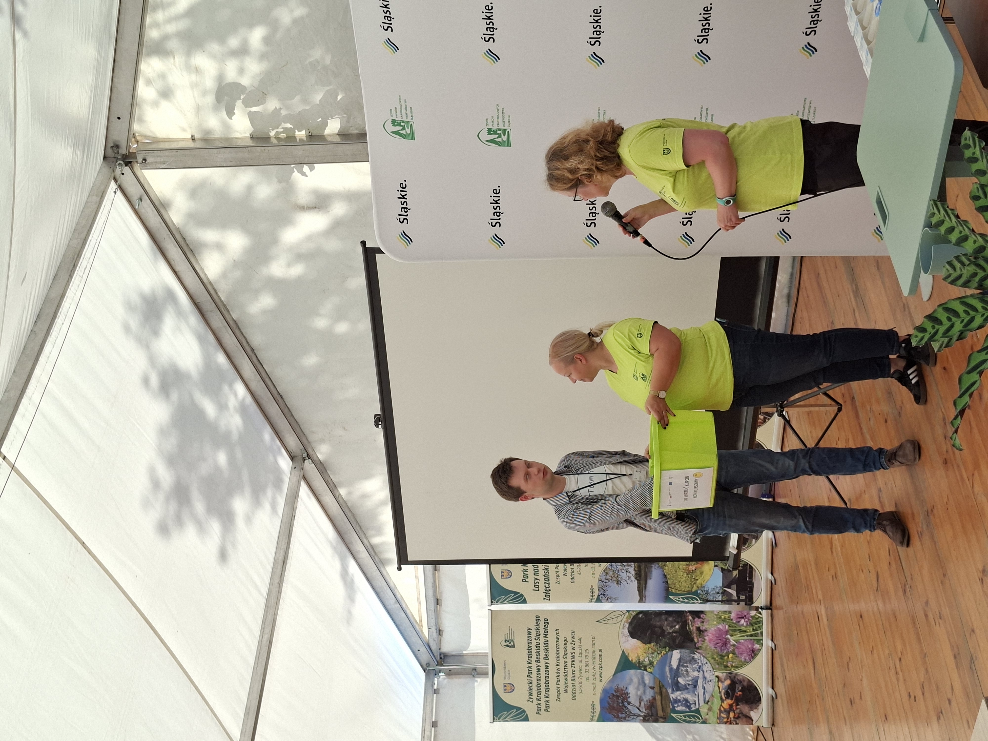 Na zdjęciu widoczne dwie uśmiechnięte kobiety ubrane w jasnozielone koszulki i ciemne spodnie  oraz wysoki mężczyzna w jasnej koszulce i niebieskiej koszuli. Wszyscy stoją na tle białego ekranu do wyświetlania prezentacji multimedialnych. Kobieta w okularach, stojąca po prawej stronie trzyma mikrofon w prawej dłoni. Kobieta na środku zdjęcia trzyma jasnozielone, plastikowe pudło, w którym mężczyzna trzyma prawą rękę.