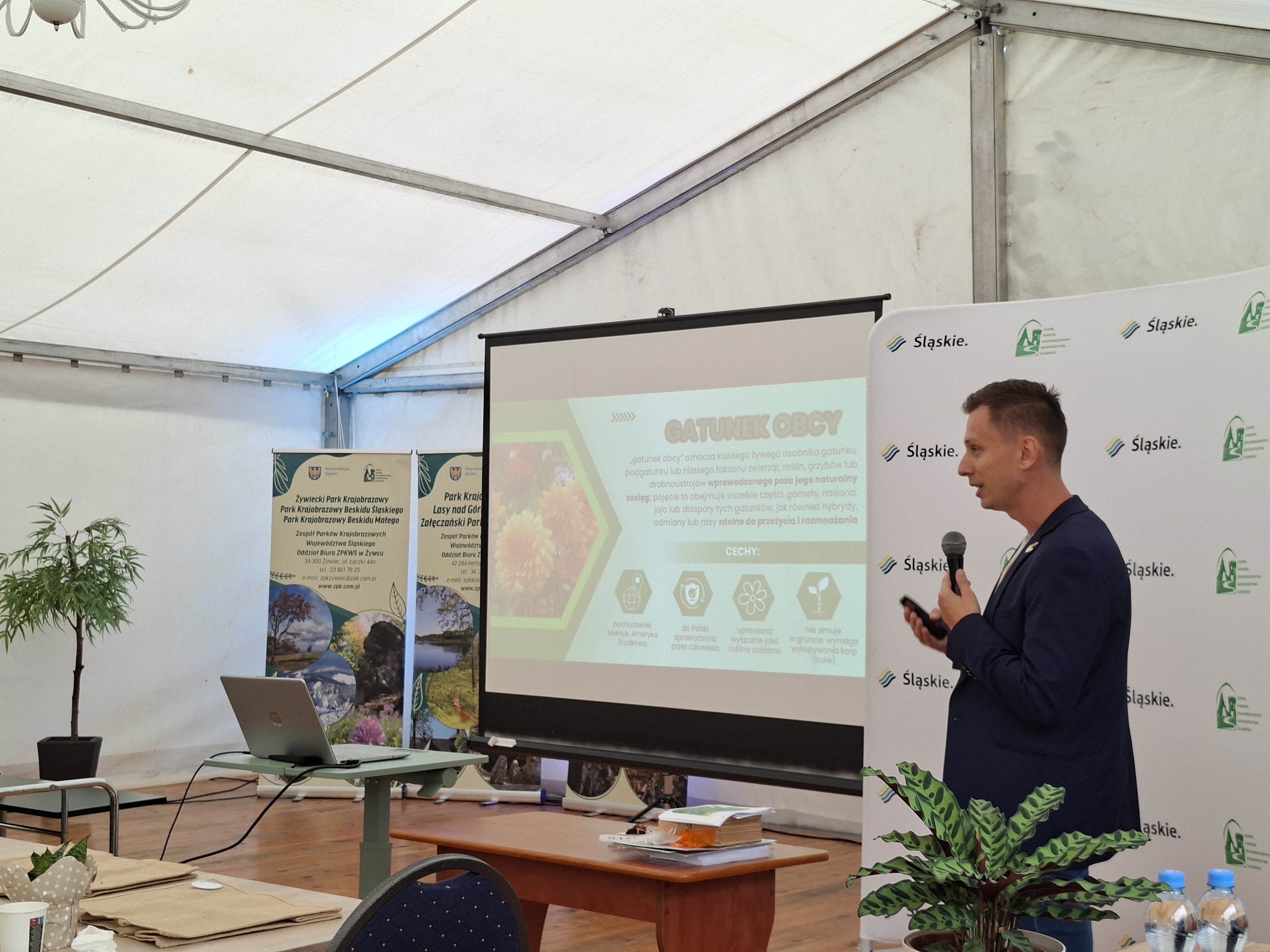 Po prawej stronie stoi mężczyzna z ciemnymi włosami i czarnej marynarce. W dłoni trzyma mikrofon. W środkowej części zdjęcia widoczny ekran z wyświetloną prezentacją multimedialną.  W lewej części zdjęcia (na drugim planie) widoczne banery informacyjne o parkach krajobrazowych.