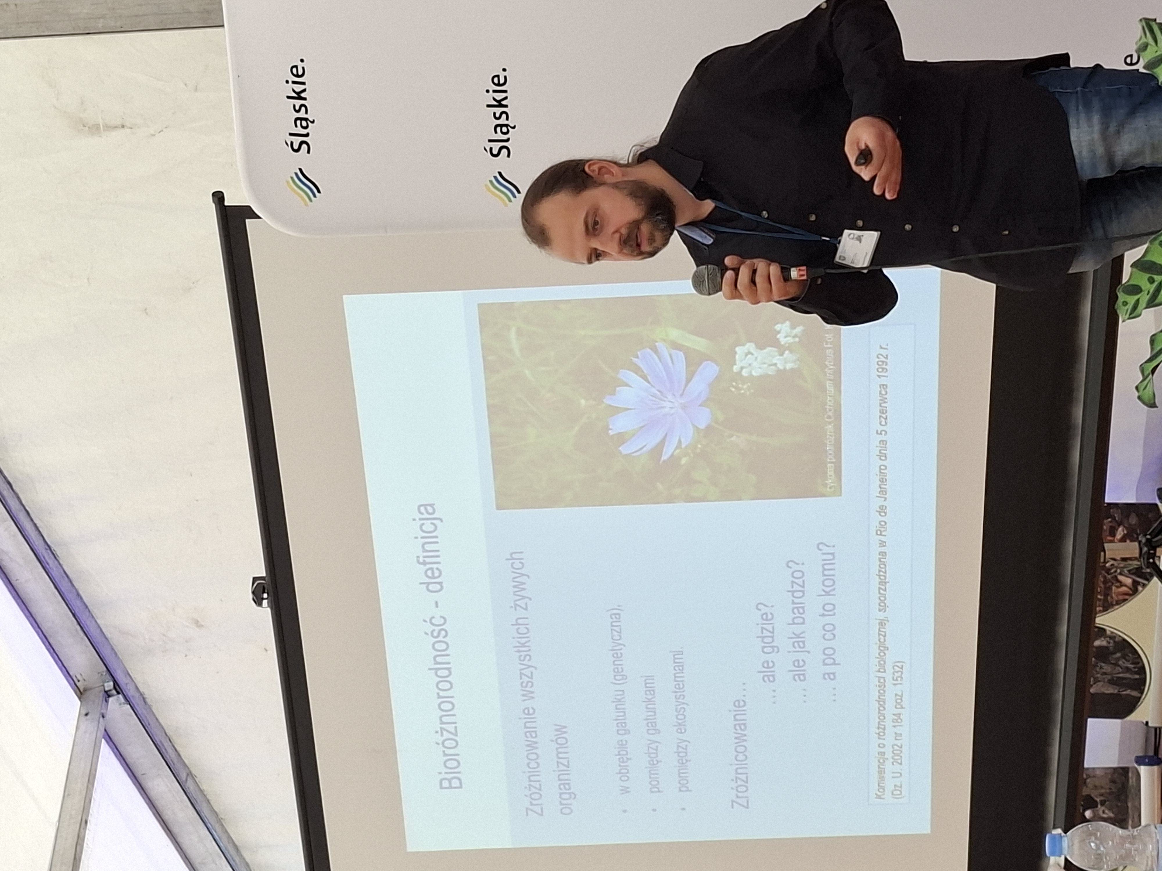 W prawym dolnym rogu zdjęcia widoczny ciemnowłosy mężczyzna z ciemnym zarostem na brodzie. Ubrany w czarną marynarkę. W prawej dłoni trzyma mikrofon. W tle widoczny ekran z wyświetloną prezentacją multimedialną.