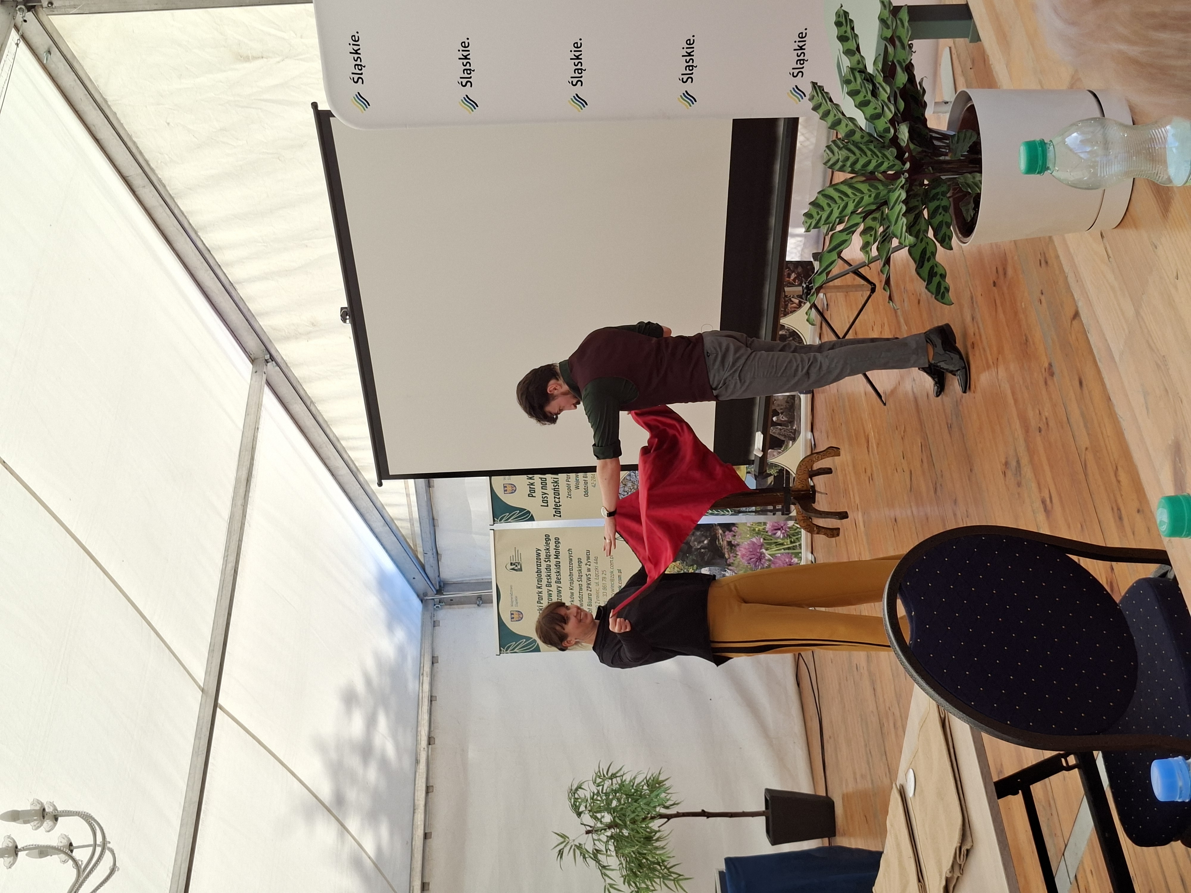 Na zdjęciu widoczna para osób - kobieta i mężczyzna. Ciemnowłosa kobieta ubrana w ciemną bluzę i jasnobrązowe spodnie, mężczyzna ubrany w brązową kamizelkę, ciemnozieloną koszulę i szare spodnie. Kobieta i mężczyzna, skierowani twarzami do siebie, trzymają za rogi czerwony obrus, pod którym znajduje się brązowy stolik.