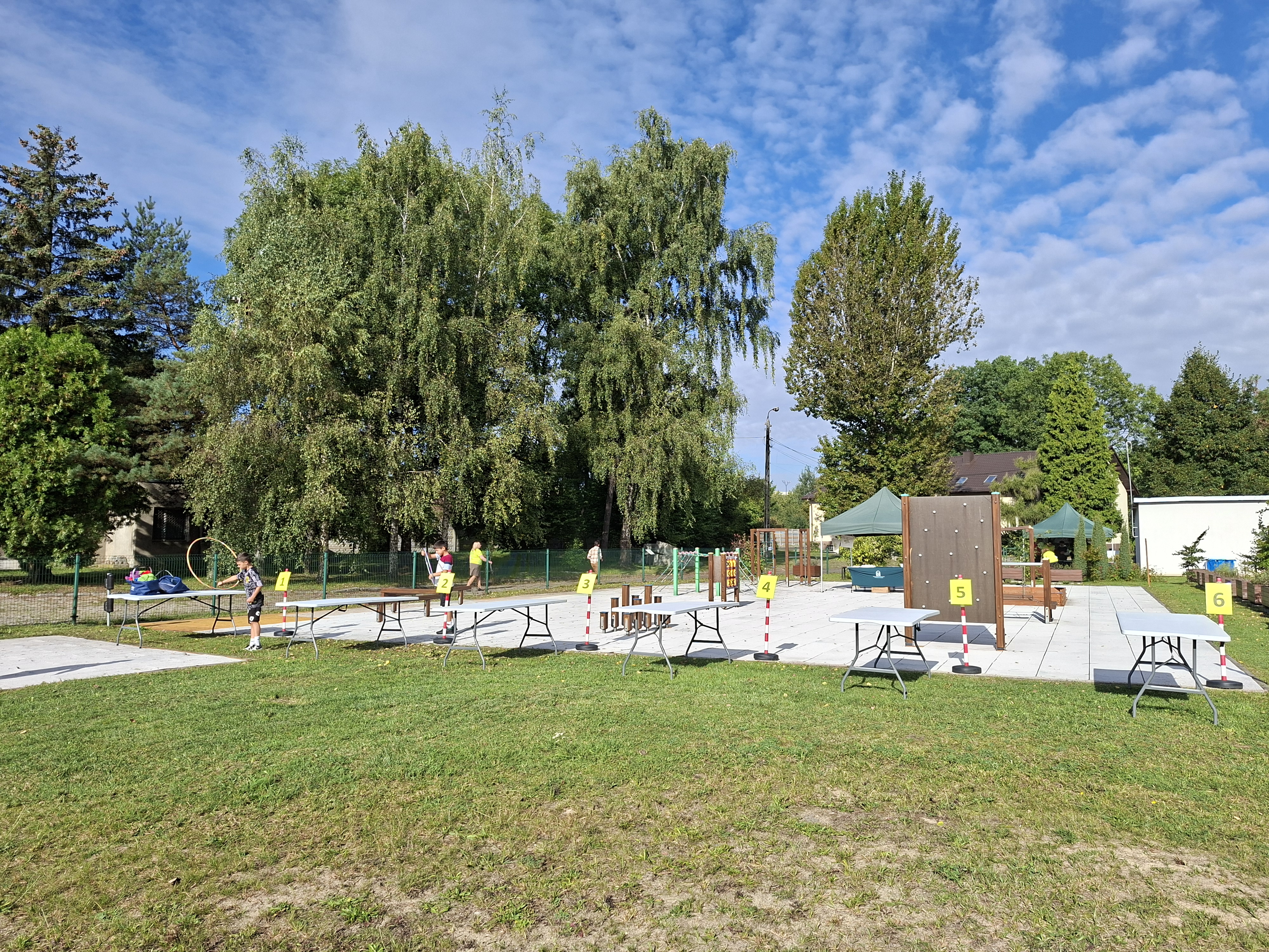 Na zdjęciu widoczny plac zabaw z urządzeniami do zabawy. Przed placem zabaw stoi sześć białych stołów. Z lewej strony chłopiec w ciemnym ubraniu bawi się hula-hop. W tle widoczne zielone drzewa liściaste i błękitne niebo.