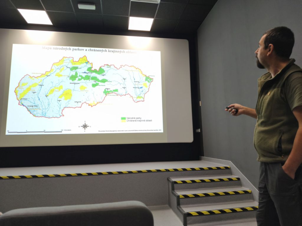 Wystąpienia Dyrektora Národný park Slovenský kras. Młody mężczyzna, wysoki, ciemne włosy i broda, ubrany w zieloną kamizelką, zielone spodnie wskaźnikiem laserowym na ekranie kinowym pokazuje obszar zarządzania parkiem. 