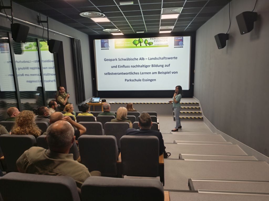 Pracownik Geopark Schwäbische Alb, kobiet w średnim wieku, ubrana w niebieską koszulką i jasne jeansy, przodem do słuchaczy siedzących na fotelach w sali kinowej rozpoczyna wygłaszać swoją prezentację. 