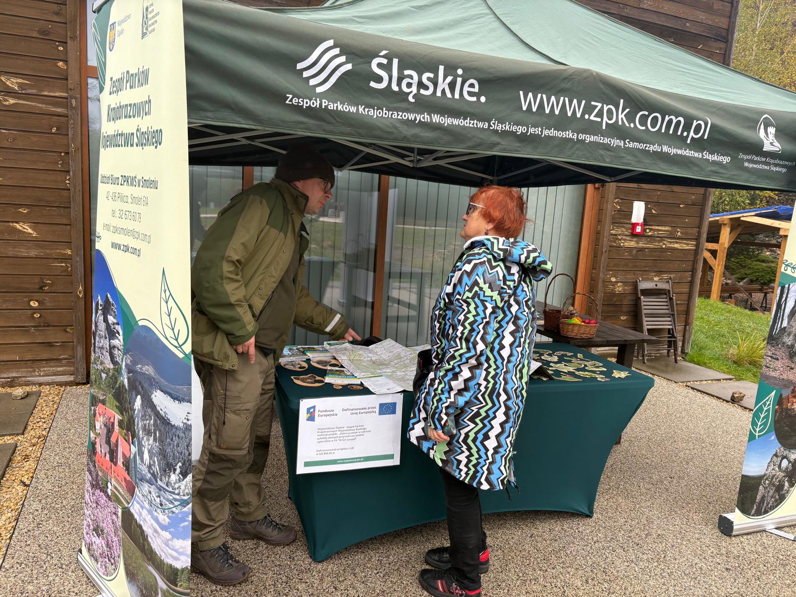 Na zdjęciu widać stoisko Zespołu Parków Krajobrazowych Województwa Śląskiego podczas Dnia Georóżnorodności w Ośrodku Edukacji Ekologiczno-Geologicznej GEOSFERA w Jaworznie. Pod zielonym namiotem z logo „Śląskie” i adresem strony internetowej www.zpk.com.pl znajduje się stół z materiałami edukacyjnymi, mapami i gadżetami promującymi przyrodę regionu. Po lewej stronie widoczny jest baner informacyjny ZPKWŚ, a obok dwoje uczestników rozmawia o prezentowanych materiałach. W tle widać drewniane budynki i fragment terenu ośrodka.
