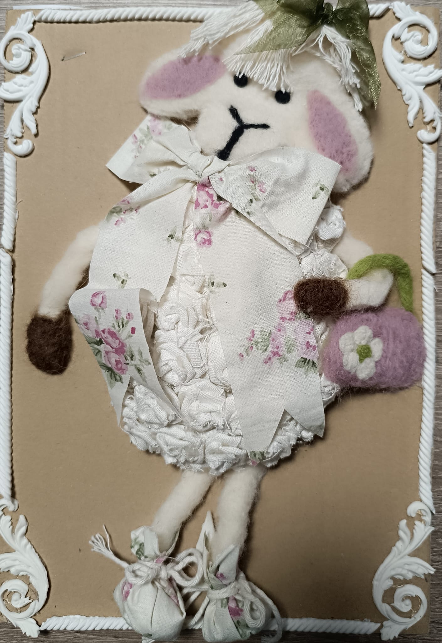 Na zdjęciu znajduje się dekoracyjna figurka owieczki wykonana z miękkich materiałów. Owieczka ma białe futerko z filcu, różowe uszy i czarne, wyszywane oczy oraz pyszczek. Ubrana jest w ozdobną sukienkę z białego materiału w kwiatowy wzór i duży kokardowy kołnierz. Na głowie ma kapelusik ozdobiony zieloną wstążką, a w łapce trzyma fioletową torebkę z białym kwiatkiem. Na nogach ma małe buciki zawiązane na kokardki. Całość umieszczona jest na beżowym tle ozdobionym białymi, dekoracyjnymi narożnikami w stylu ramki.