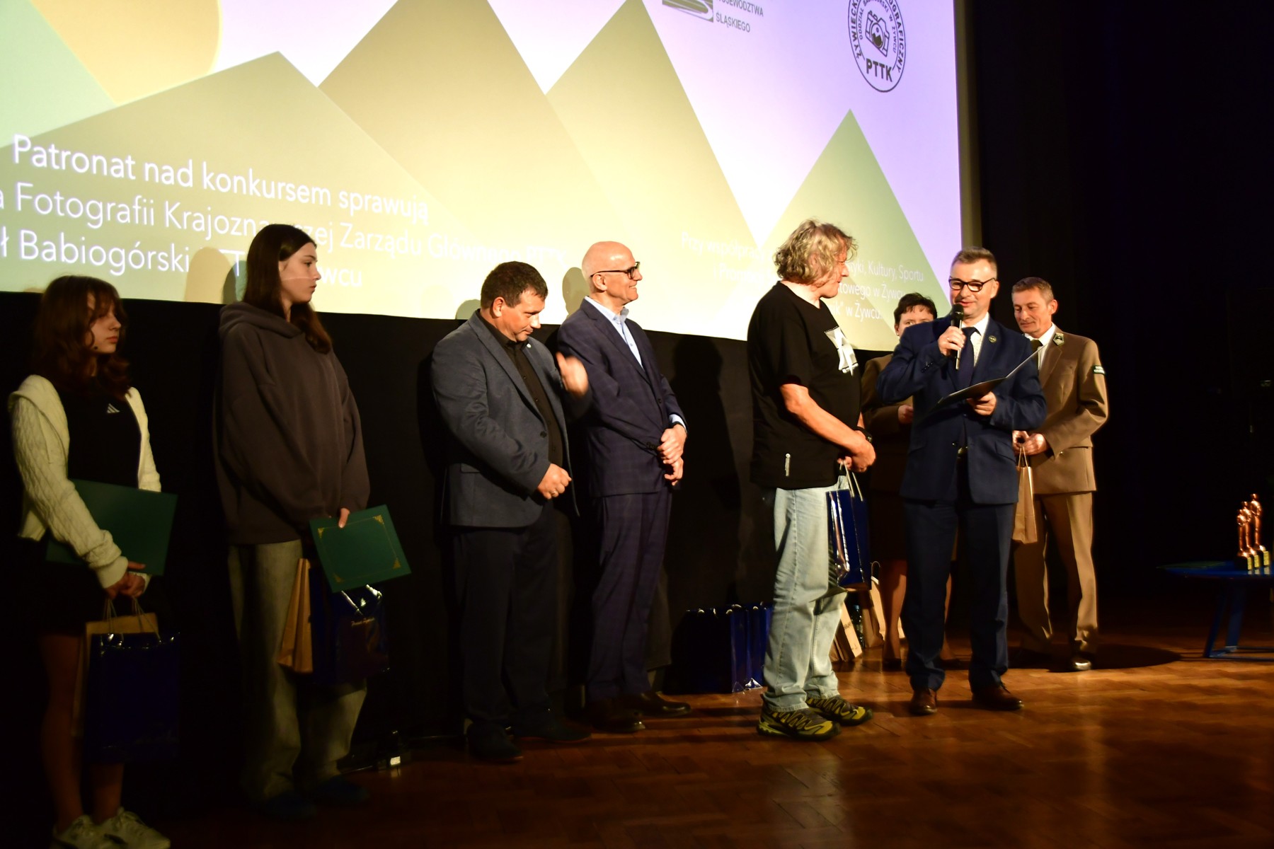 Organizatory i laureaci konkursu stoją na scenie. Jeden z prowadzących przemawia do zgromadzonych.