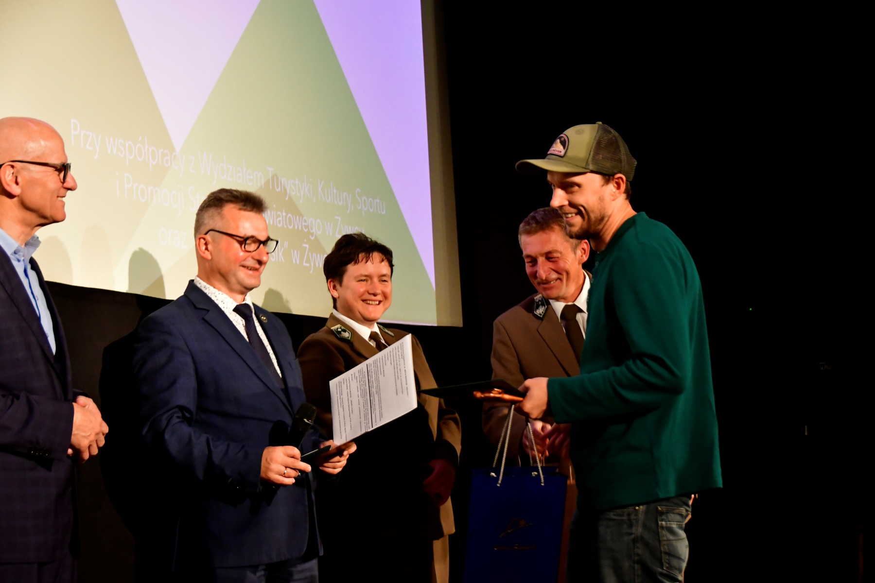 Prowadzący wręcza nagrodę uczestnikowi konkursu. W otoczeniu pozostali organizatorzy konkursu.