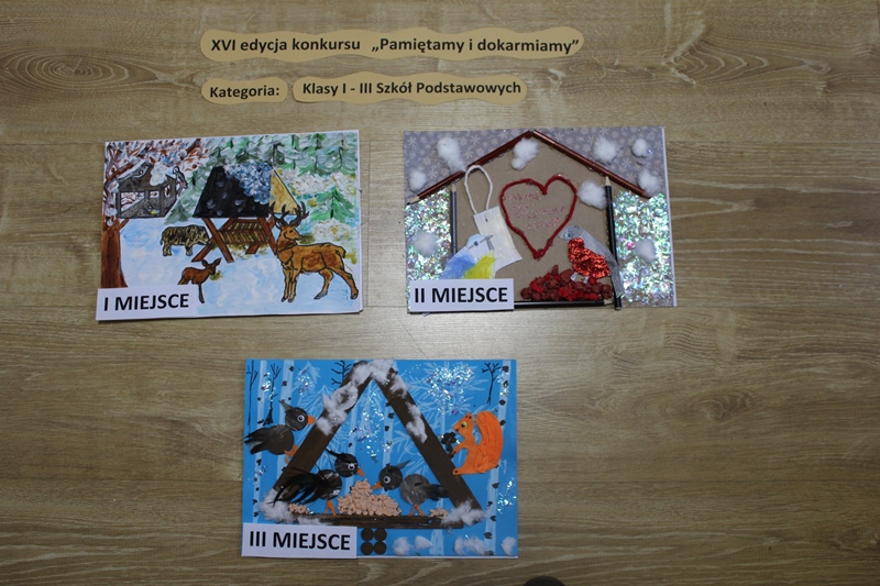 Zdjęcie przedstawia prace laureatów konkursu w kategorii klasy I-III szkoły podstawowej. I miejsce namalowane zwierzęta w lesie przy paśniku. II miejsce praca przedstawia karmnik z kredek, a w nim ptaki z przyklejonymi piórami oraz owoce z bibuły. III miejsce karmnik z patyczków oraz ptaki z drewna z pórami.