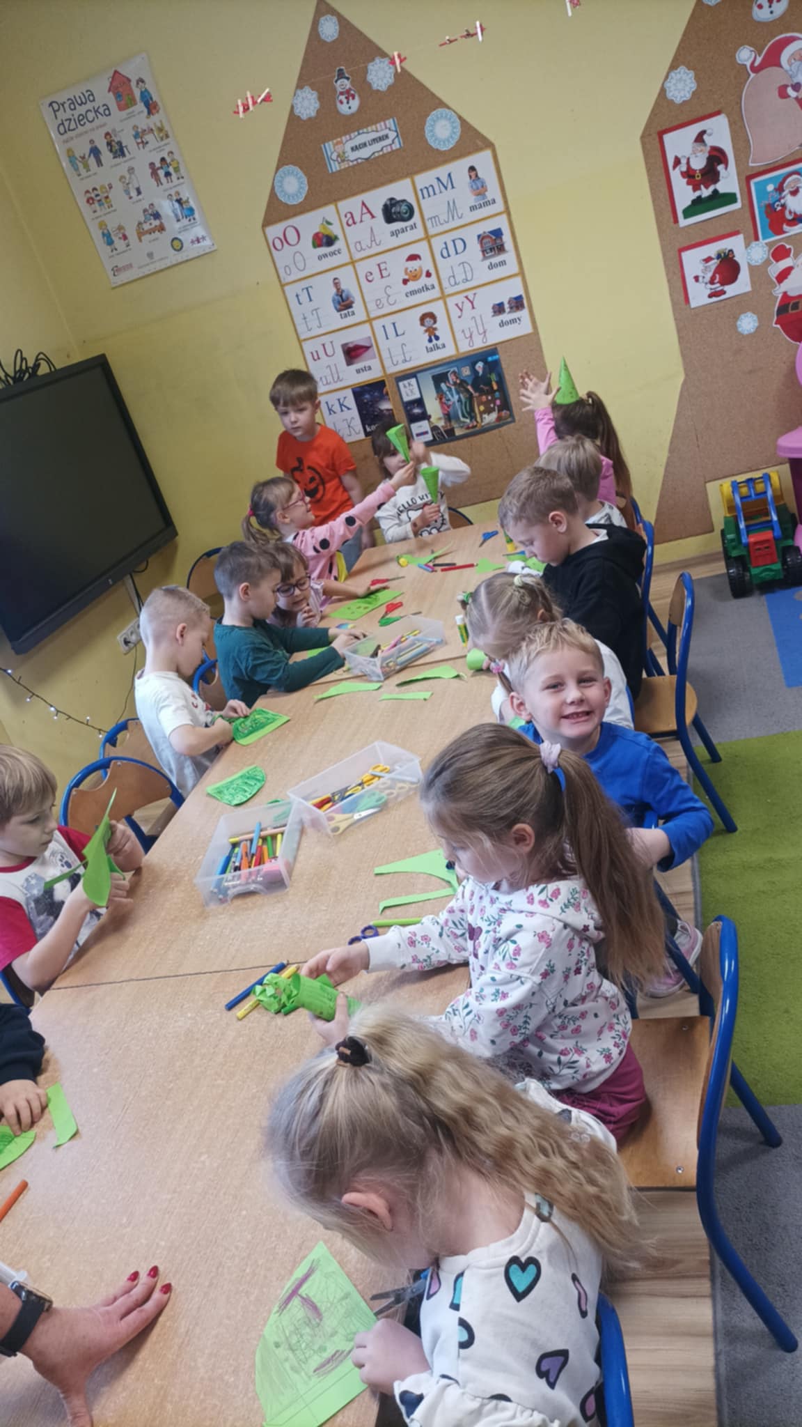 Przedszkolaki siedzą przy stole i wykonują choinkę z papieru.