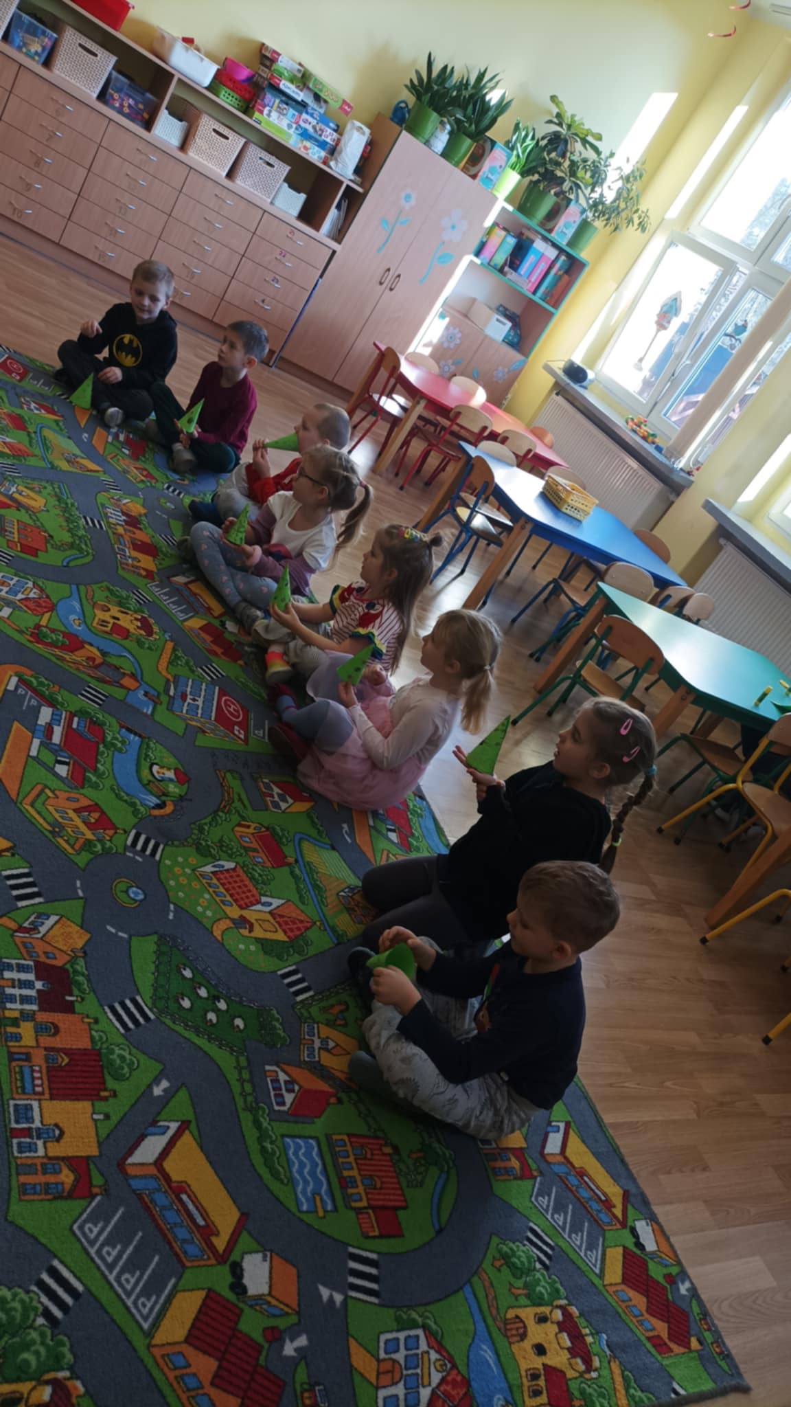 Przedszkolaki siedzą na dywanie i trzymają gotowe choinki z papieru.