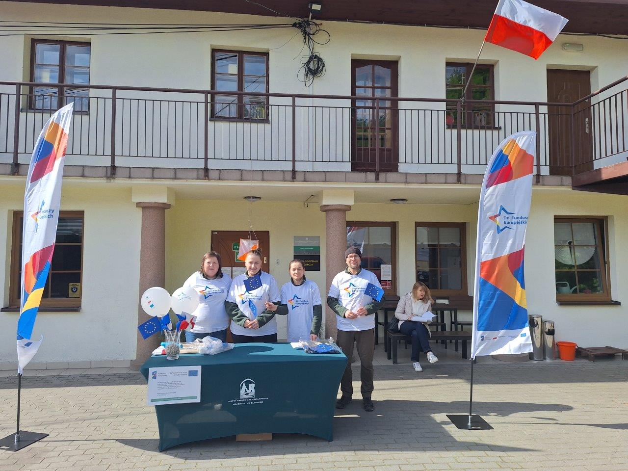 widać biuro oddziału ZPKWŚ w Smoleniu, przed biurem stoją pracownicy ZPKWŚ ubrani w koszulki z logotypami Dni Funduszy Europejskich. Na stole w wazonie są flagi Unii Europejskiej, Polski oraz balony z logotypami. Na stole zamieszczony jest plakat dot. realizacji projektu przez ZPKWŚ pt. „Dobre praktyki w zakresie ochrony cennych przyrodniczo siedlisk i gatunków w PK ’’Orlich Gniazd” nr FENX.01.05-IW.01-0023/24 w ramach działania FENX.01.05.Ochrona przyrody i rozwój zielonej infrastruktury priorytetu FENX.01 Wsparcie sektorów energetyka i środowisko z Funduszu Spójności. Obok stołu stoją dwa windery (flagi) z logotypem Dni Funduszy Europejskich.
