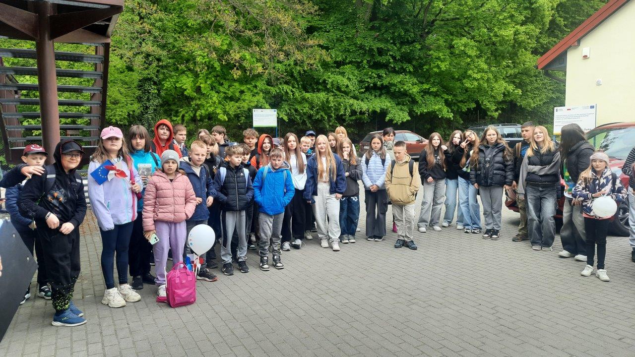 widać grupę dzieci stojących przed budynkiem ZPKWŚ w Smoleniu, dzieci są uśmiechnięte, trzymają w rękach balony i flagi Unii Europejskiej i Polski. W oddali widać las.  