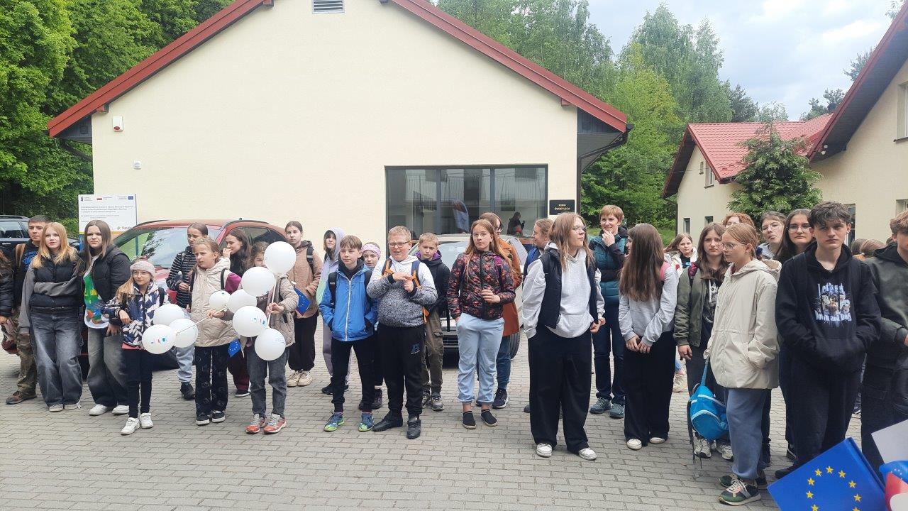 widać grupę dzieci stojących przed budynkiem ZPKWŚ w Smoleniu, dwie dziewczynki trzymają balony z logotypami Unii Europejskiej i Polski. Twarze dzieci wskazują, iż są one zadowolone. Na drugim planie widać budynek świetlicy – kino oraz część budynków Jurajskiej Ostoi.  