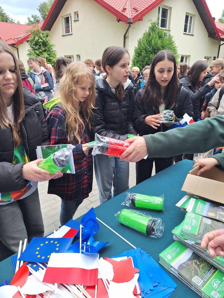 widać dzieci odbierające nagrody bidony książki i mapy, ich twarze są uśmiechnięte. Na stole widać leżące flagi Unii Europejskiej i Polski oraz bidony, książki i mapy. W oddali widać grupę dzieci oraz ośrodek Jurajskiej Ostoi. 