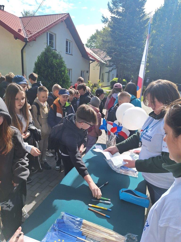 widać pracowników ZPKWŚ oddziału w Smoleniu, którzy rozdają karty startowe dzieciom do wypisania nazwy grupy, nazwiska oraz inne. Dzieci z ciekawością podchodzą do stołu i odbierają karty. 