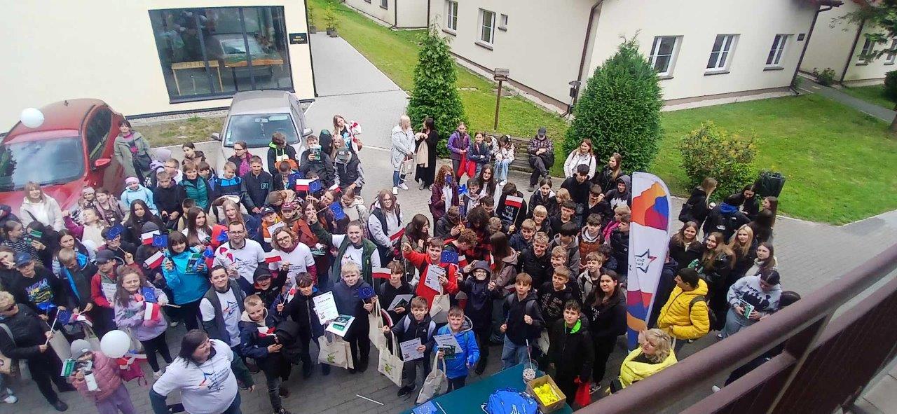 widać dużą grupę dzieci z nauczycielami oraz pracowników ZPKWŚ. Widać że są uśmiechnięci. W rękach trzymają balony flagi z logotypami Unii Europejskiej i Polski. W oddali widać budynki ZPKWŚ. 