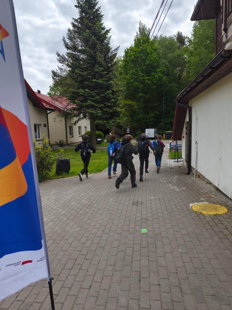 widać dzieci rozpoczynające grę, biegną w kierunku, którym mapa im wskazuje. W rękach trzymają flagę Unii Europejskiej i Polski. W oddali widać budynek Jurajskiej Ostoi oraz duże drzewa. 