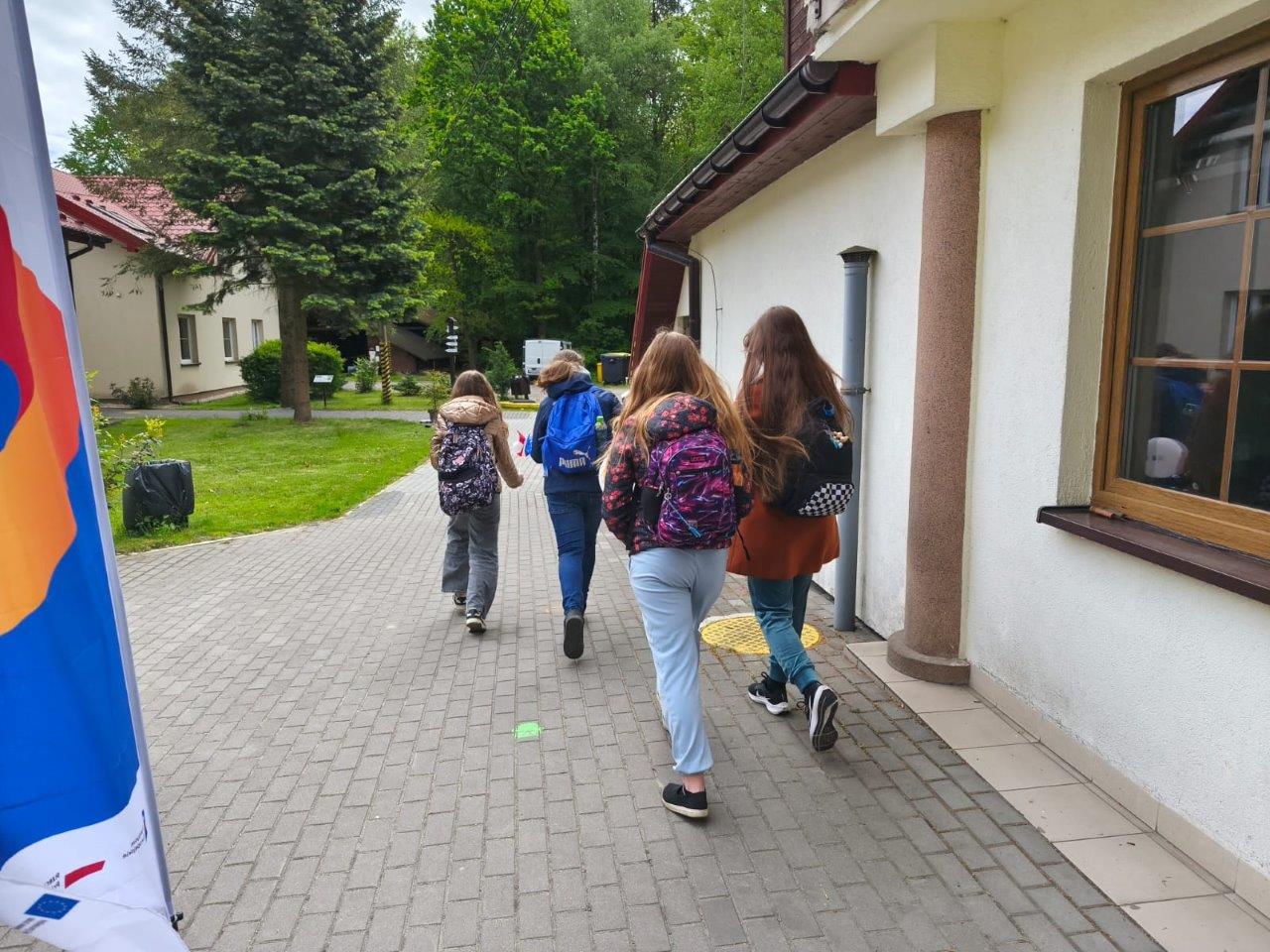widać dzieci rozpoczynające grę, biegną w kierunku, którym mapa im wskazuje. W rękach trzymają flagę Unii Europejskiej i Polski. W oddali widać budynek Jurajskiej Ostoi oraz duże drzewa. 