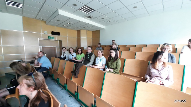 Grupa uczestników siedzi w sali wykładowej na drewnianych krzesłach audytoryjnych. Pomieszczenie jest jasne, z białymi ścianami, drewnianą boazerią i podwieszanym sufitem. W tle widoczny jest zielony znak wyjścia ewakuacyjnego oraz drzwi. Uczestnicy skupiają się na prezentacji, niektórzy trzymają butelki wody.