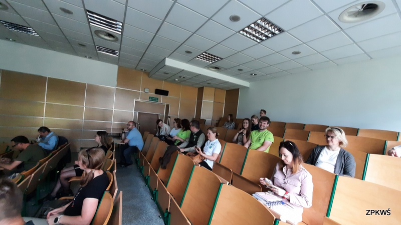 Grupa uczestników siedzi w sali wykładowej na drewnianych krzesłach audytoryjnych. Pomieszczenie jest jasne, z białymi ścianami, drewnianą boazerią i podwieszanym sufitem. W tle widoczny jest zielony znak wyjścia ewakuacyjnego oraz drzwi. Uczestnicy skupiają się na prezentacji, niektórzy trzymają telefony w ręce. 