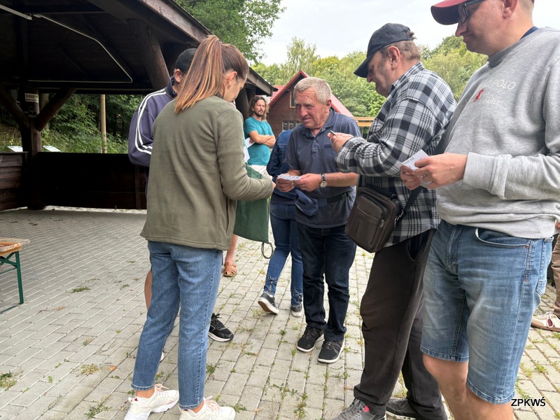 Na zdjęciu widać grupę osób zgromadzonych na utwardzonym placu wyłożonym kostką brukową, obok drewnianej wiaty. Ludzie stoją blisko siebie i trzymają w rękach kartki papieru, lub karty do gry. Kobieta w zielonej kurtce i dżinsach przekazuje coś mężczyźnie w starszym wieku, ubranym w granatową kurtkę. Pozostali uczestnicy skupiają się na swoich kartkach, jakby porównywali je lub sprawdzali. W tle widać kolejne osoby stojące i obserwujące sytuację, a także fragmenty drewnianych zabudowań oraz zieleń drzew.