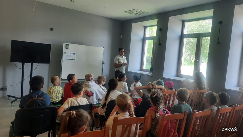 Grupa dzieci siedzących na krzesłach podczas warsztatów edukacyjnych w sali z dużymi oknami, słuchających prezentacji prowadzonej przez instruktora. W tle widać tablicę na której zamieszczony jest plakat informacyjno-promocyjny dot. „Dobre praktyki w zakresie ochrony cennych przyrodniczo siedlisk i gatunków w PK ’’Orlich Gniazd” nr FENX.01.05-IW.01-0023/24 oraz logotypy Funduszy Europejskich i Unii Europejskiej, duży monitor, a za oknami zielony krajobraz.