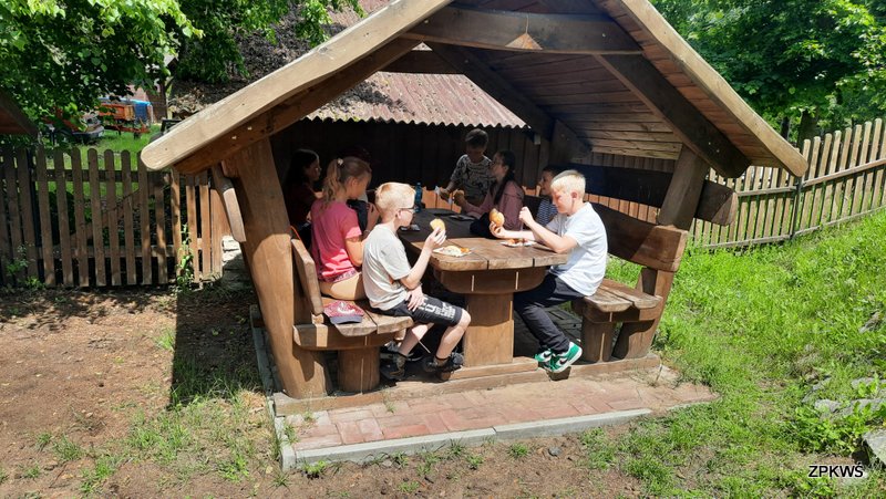 Grupa dzieci siedzi na ławkach w altance, która stoi na podeście z kostki brukowej. Na stoliku są talerzyki papierowe a na nich kiełbaski, ketchup i bułki. Dwóch chłopców w ręce trzymają bułkę. Altanka jest drewniana, w tle widać szopę pokrytą blachą w kształcie dachówki oraz trawę. 