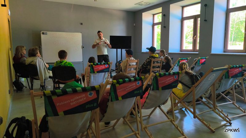 Grupa dzieci i opiekunów siedzących na leżakach i krzesłach podczas warsztatów edukacyjnych. Na środku sali stoi instruktor GOPR, prezentujący treści związane z bezpieczeństwem na jurajskich szlakach. W tle widać tablicę i duży ekran, a za oknami zielony krajobraz