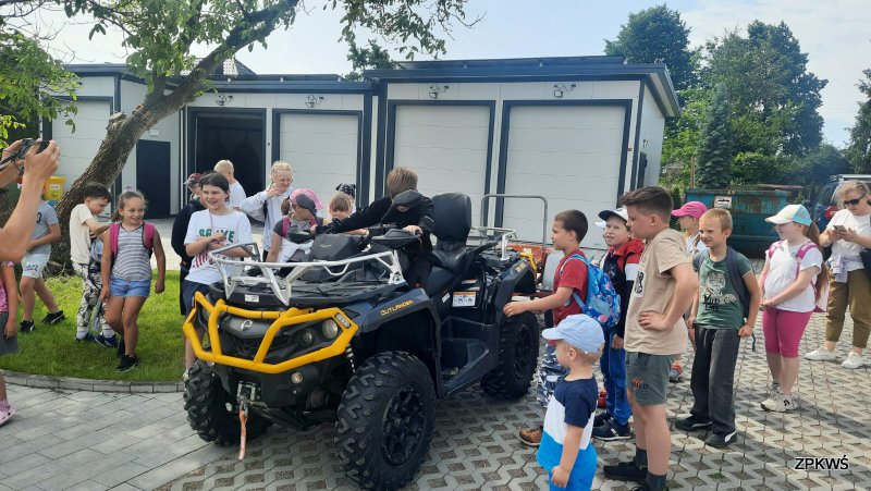 Grupa dzieci w różnym wieku stoi na brukowanym placu wokół dużego, czarno-żółtego quada ratowników GOPR. Część dzieci znajduje się bezpośrednio przy pojeździe – jedno siedzi na quadzie, inne spoglądają zaciekawione albo rozmawiają ze sobą. Widoczni są zarówno chłopcy, jak i dziewczynki, większość z plecakami, w letnich ubraniach. W tle widać jasne budynki garażowe, zielone drzewa i kawałek nieba. Atmosfera jest swobodna i edukacyjna, typowa dla spotkania dzieci ze służbami ratunkowymi.