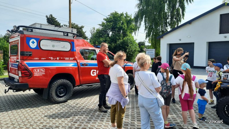 Grupa dzieci w różnym wieku, instruktor GOPR oraz opiekunowie dzieci stoją na brukowanym placu obok dużego czerwonego pojazdu. Jest to pojazd ratunkowy GOPR z niebieskimi pasami i białymi napisami GOPR. Jeden z opiekunów robi zdjęcia dzieciom stojących i słuchających instruktora GOPR. W tle widać duży biały z grafitową bramą garaż oraz zielone drzewa. 