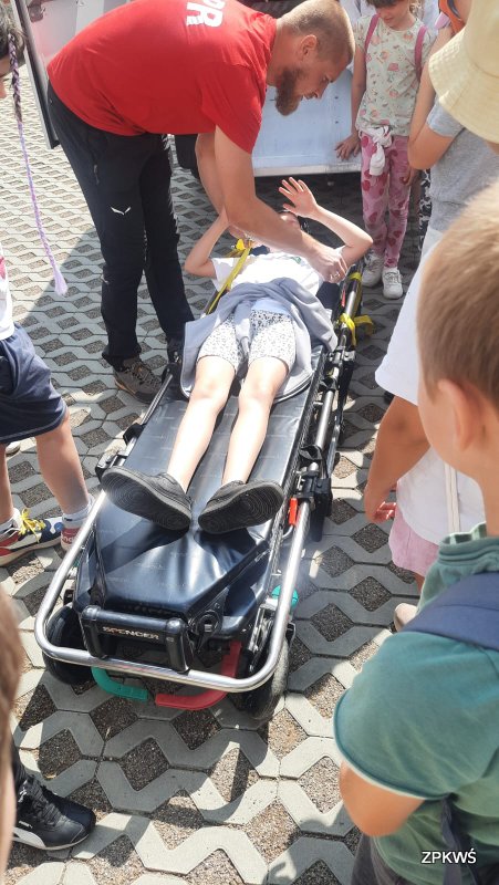 Grupa dzieci w różnym wieku stoi na brukowanym placu wokół noszy ratowniczych. Instruktor GOPR ubrany w czerwoną koszulkę jest pochylony nad noszami na których leży dziewczynka. Instruktor spina ją żółtymi pasami dla bezpieczeństwa, aby dziewczynka nie spadła podczas przewożenia ją do pojazdu ratowniczego GOPR. 