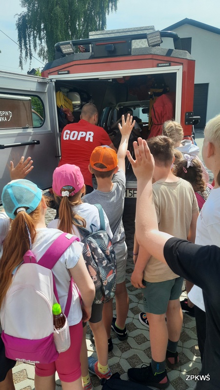 Grupa dzieci w różnym wieku stoi przed pojazdem ratowniczym GOPR, dzieci na głowie mają kolorowe czapki: pomarańczowa, różowa i niebieska. Drzwi w pojeździe GOPR są otwarte. W pojeździe na noszach znajduje się dziewczynka spięta pasami. Przed samochodem stoi instruktor GOPR i zabezpiecza nosze w pojeździe. 
