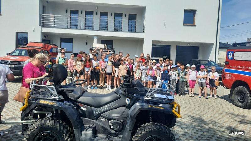 Grupa dzieci, opiekunowie oraz instruktorzy GOPR stoją na brukowanym placu ustawiają się do zdjęcia, są uśmiechnięci i rozmawiają. Całą grupa jest ubrana w różnokolorowe ubrania część grupy ma czapki założone na głowie. W tle widać duży biały budynek z dużymi oknami oraz balkon. Z prawej strony widać pojazd czerwony GOPR a przed grupą stoi czarny z żółtymi wstawkami quad GOPR. 
