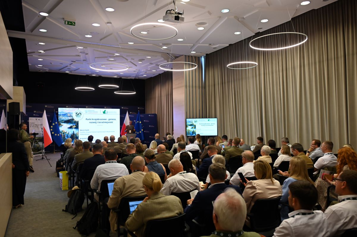 uczestnicy konferencji siedzą w sali i biorą aktywny udział w wydarzeniu. Zdjęcie perspektywiczne ujmujące wszystkich uczestników w tle znajduje się mównica i główna plansza informacyjna konferencji.